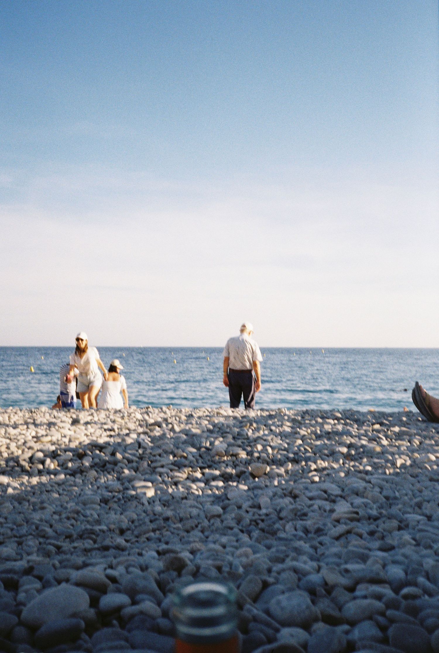 Family walking on a pebble beach toward the calm blue sea under a clear sky.