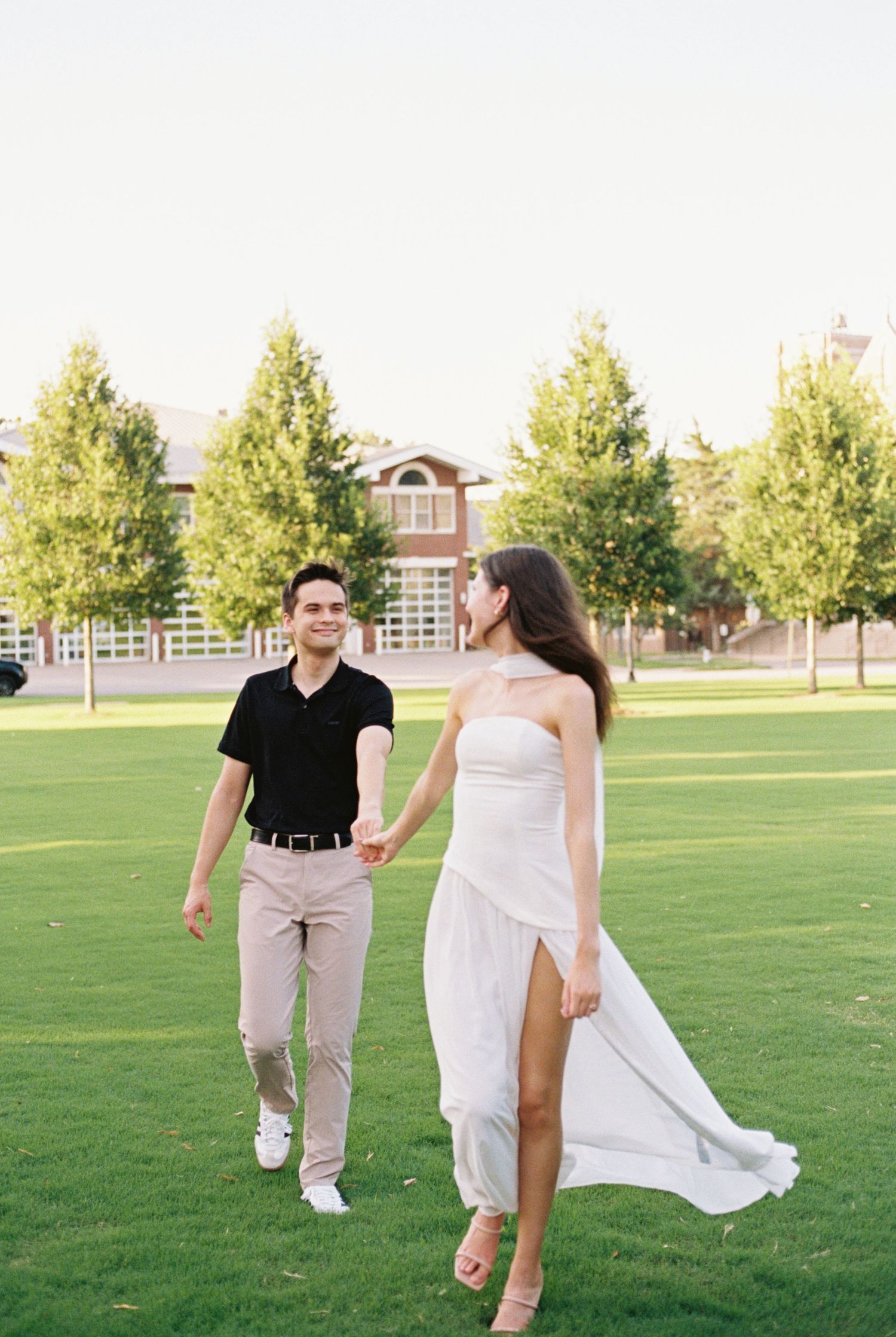 Couple holding hands walking across a green lawn during golden hour engagement photography session.