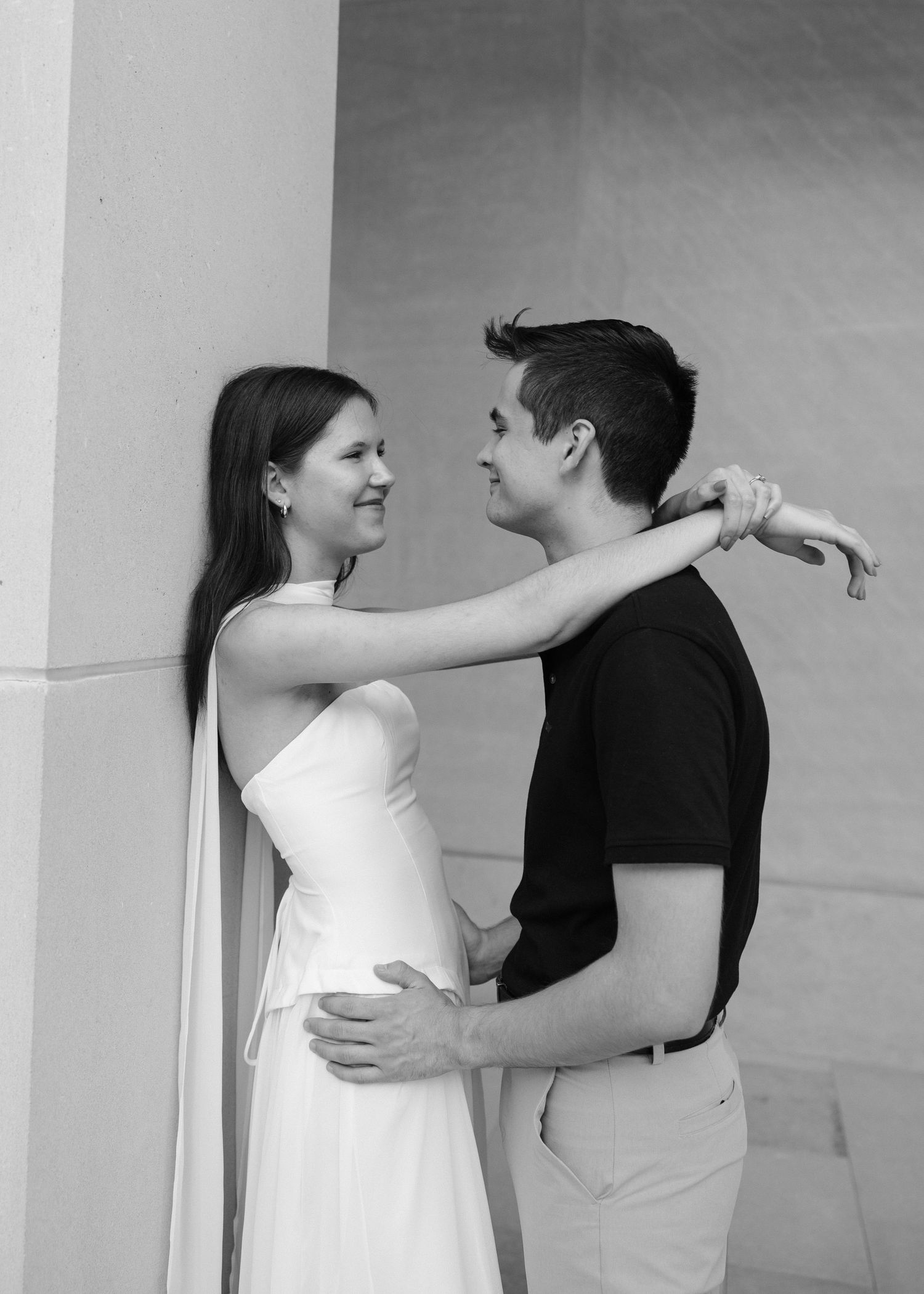 Black and white photo of a couple embracing and smiling at each other against a wall.