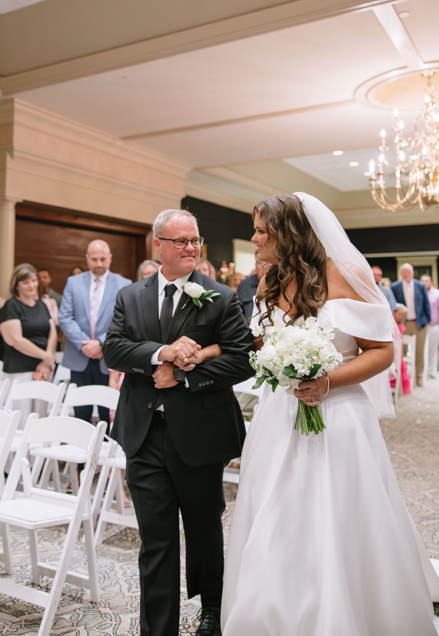 Bride in off-shoulder gown walks down aisle with father during indoor wedding ceremony.