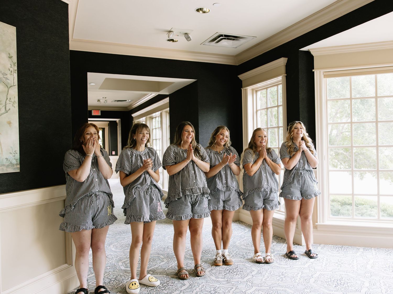 Six bridesmaids in matching gray ruffle rompers pose together in a bright elegant room before a wedding ceremony.