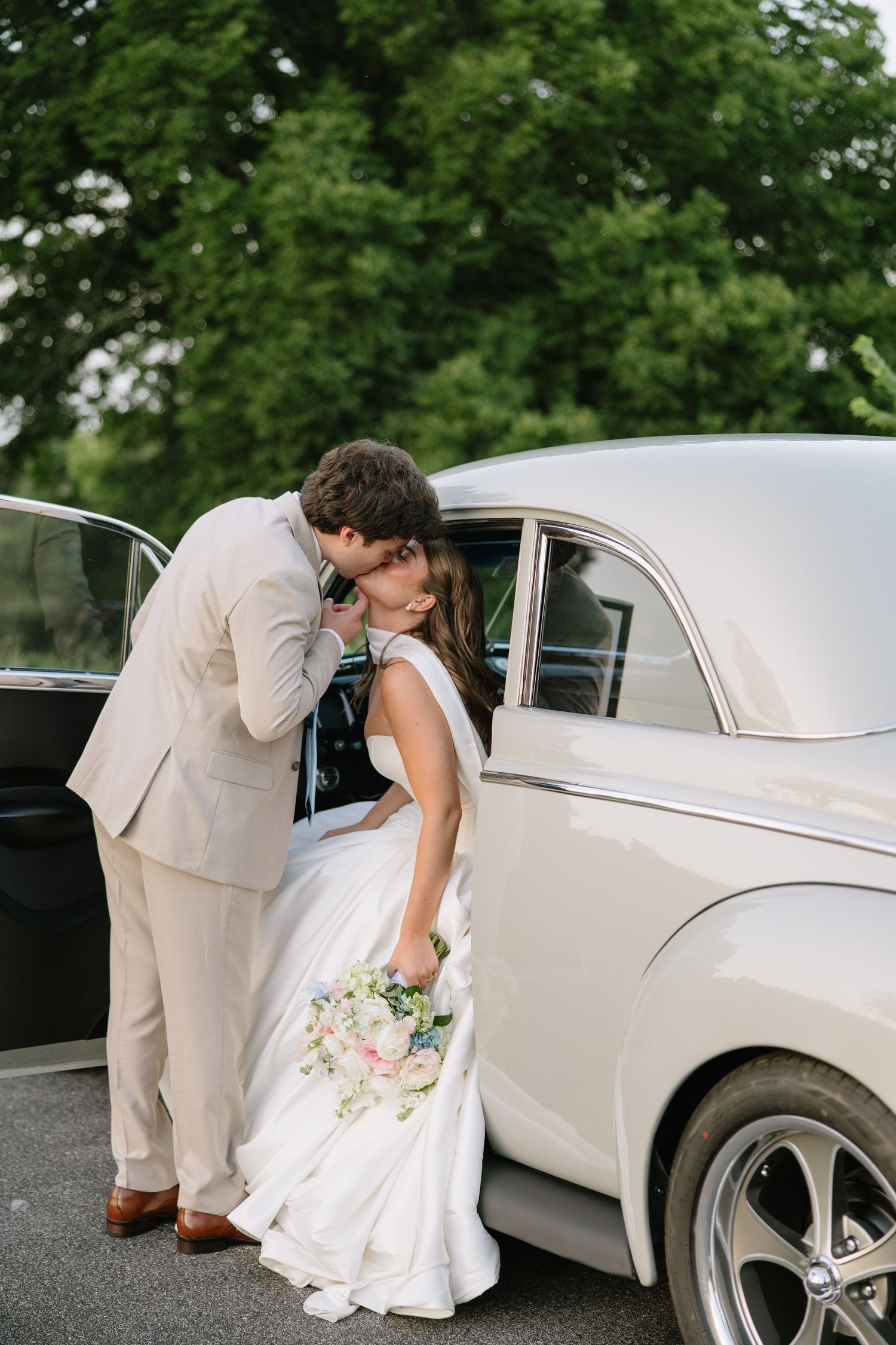 Bride and groom sharing a kiss beside a classic white vintage car, bride holding a pastel bouquet in a white gown.