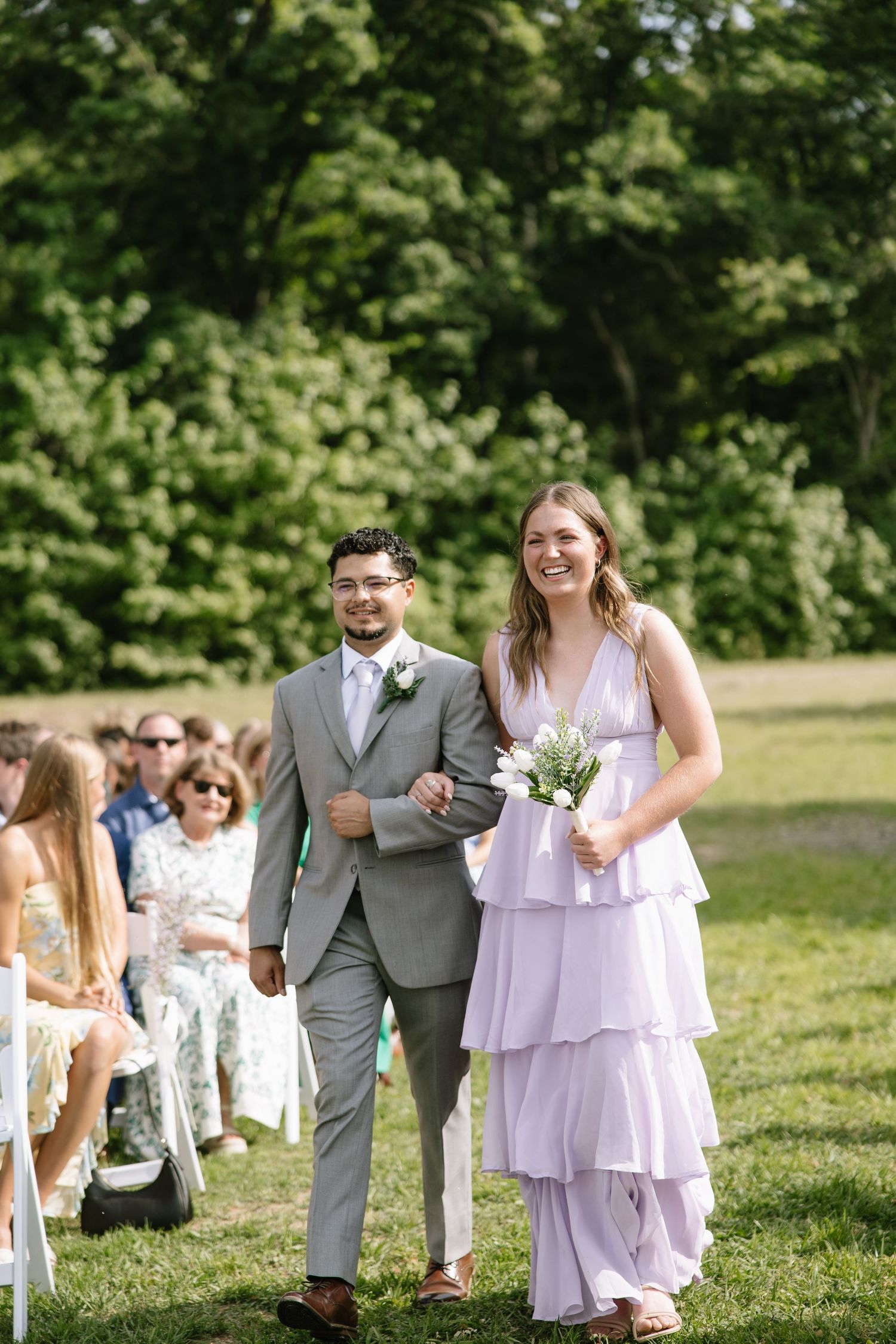 A smiling bridesmaid in lavender dress walks down outdoor aisle with groomsman in gray suit at garden wedding.