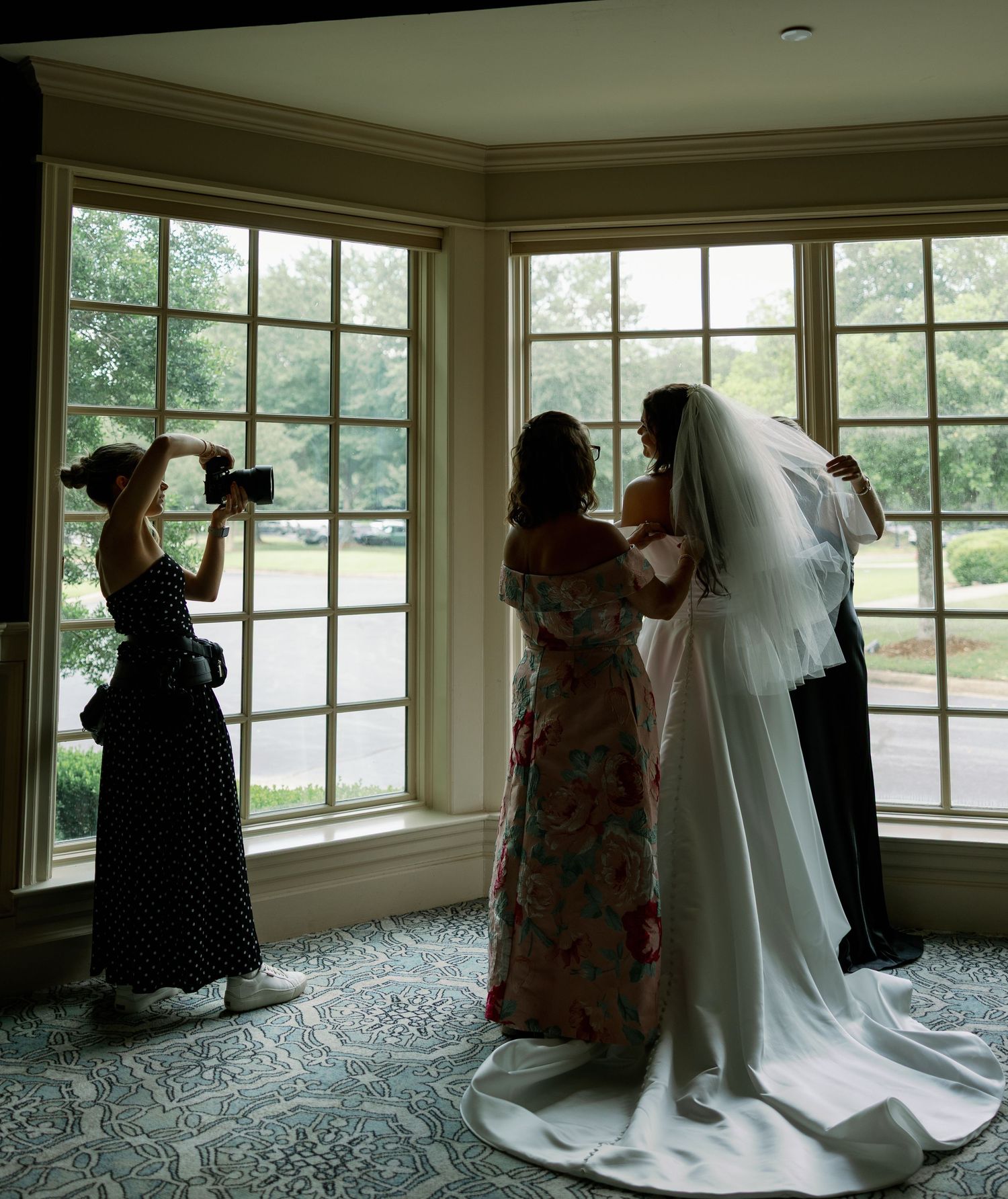 Bride in white gown and veil being assisted by guests near bay windows while photographer captures the moment.