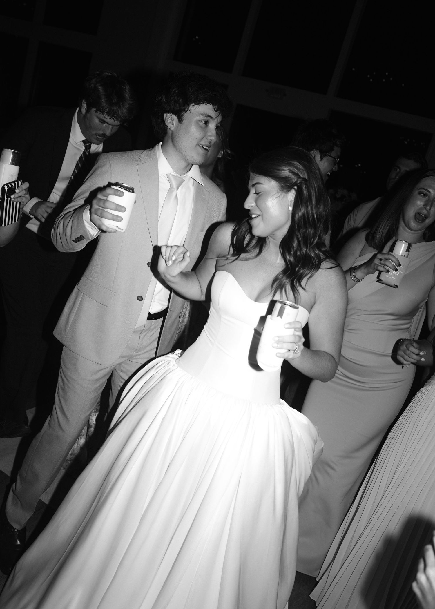 Black and white photo of a couple dancing at a wedding reception, bride in strapless gown holding drinks.