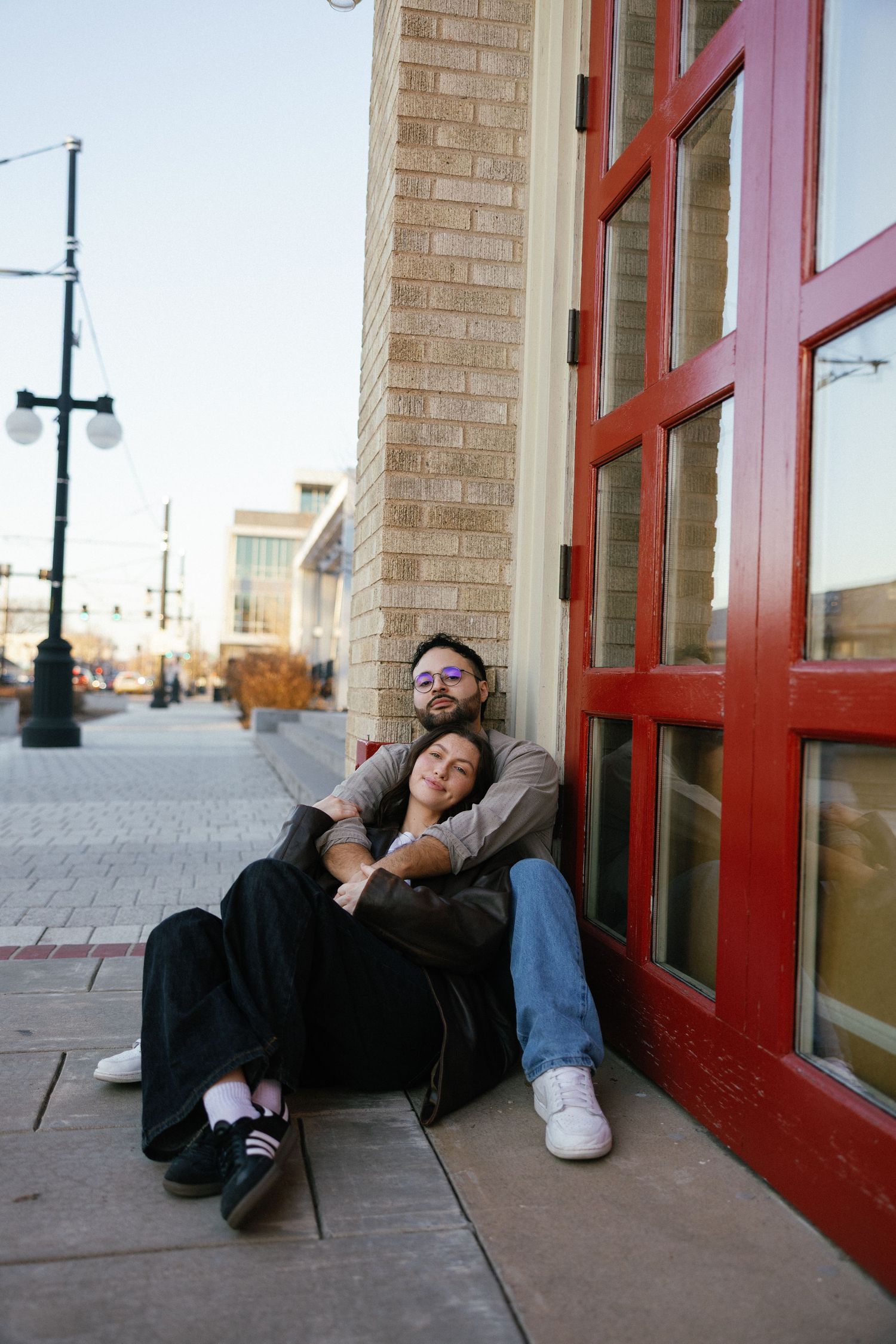 Couple sitting together on urban sidewalk against brick building with red door, woman resting head on man's shoulder.