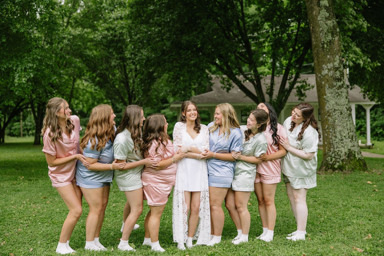 Bride in white dress surrounded by bridesmaids in colorful matching outfits hugging outdoors on green lawn.