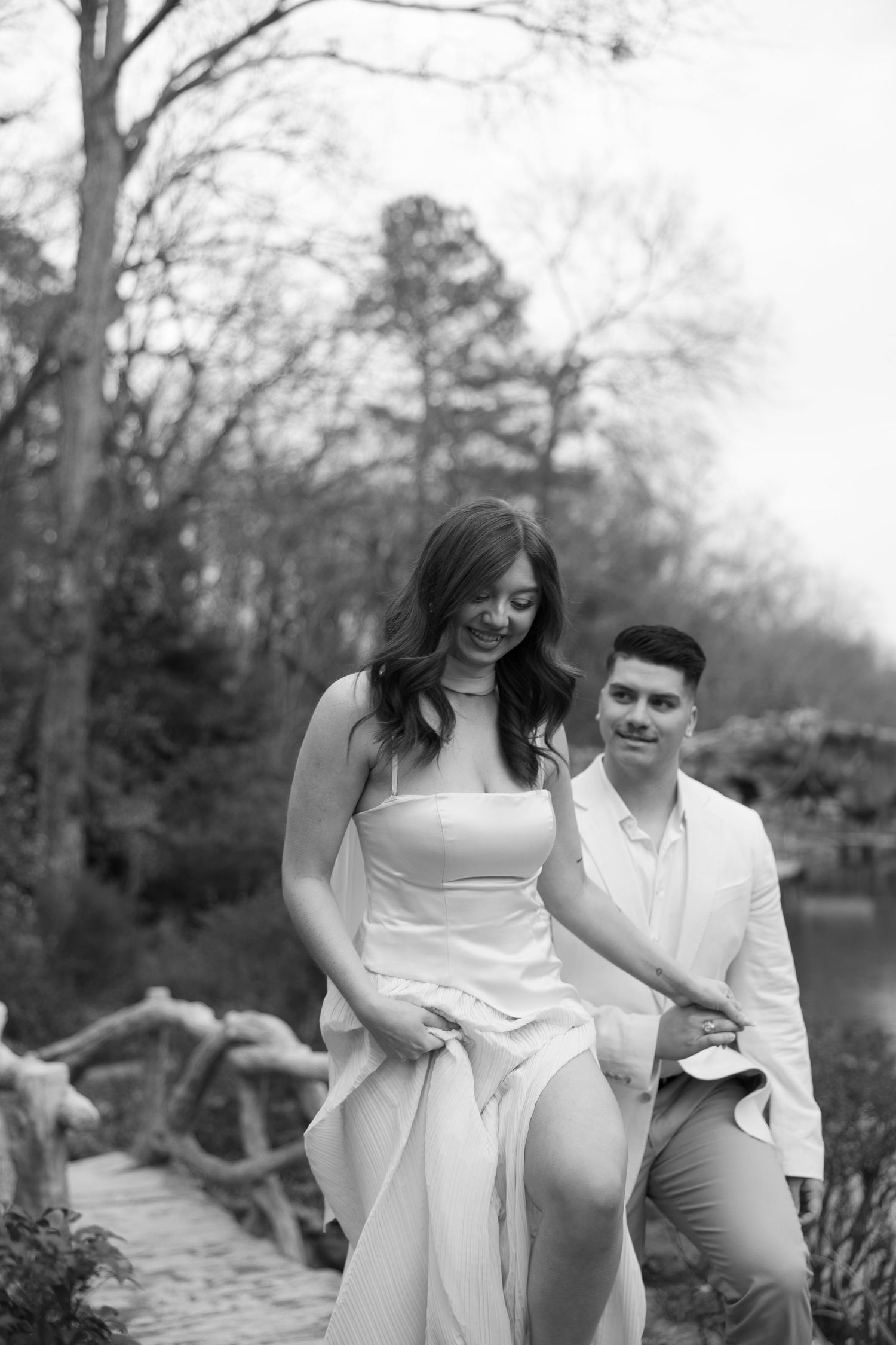 Black and white photo of a couple walking on a rustic bridge, man in white suit smiling at woman in elegant dress.