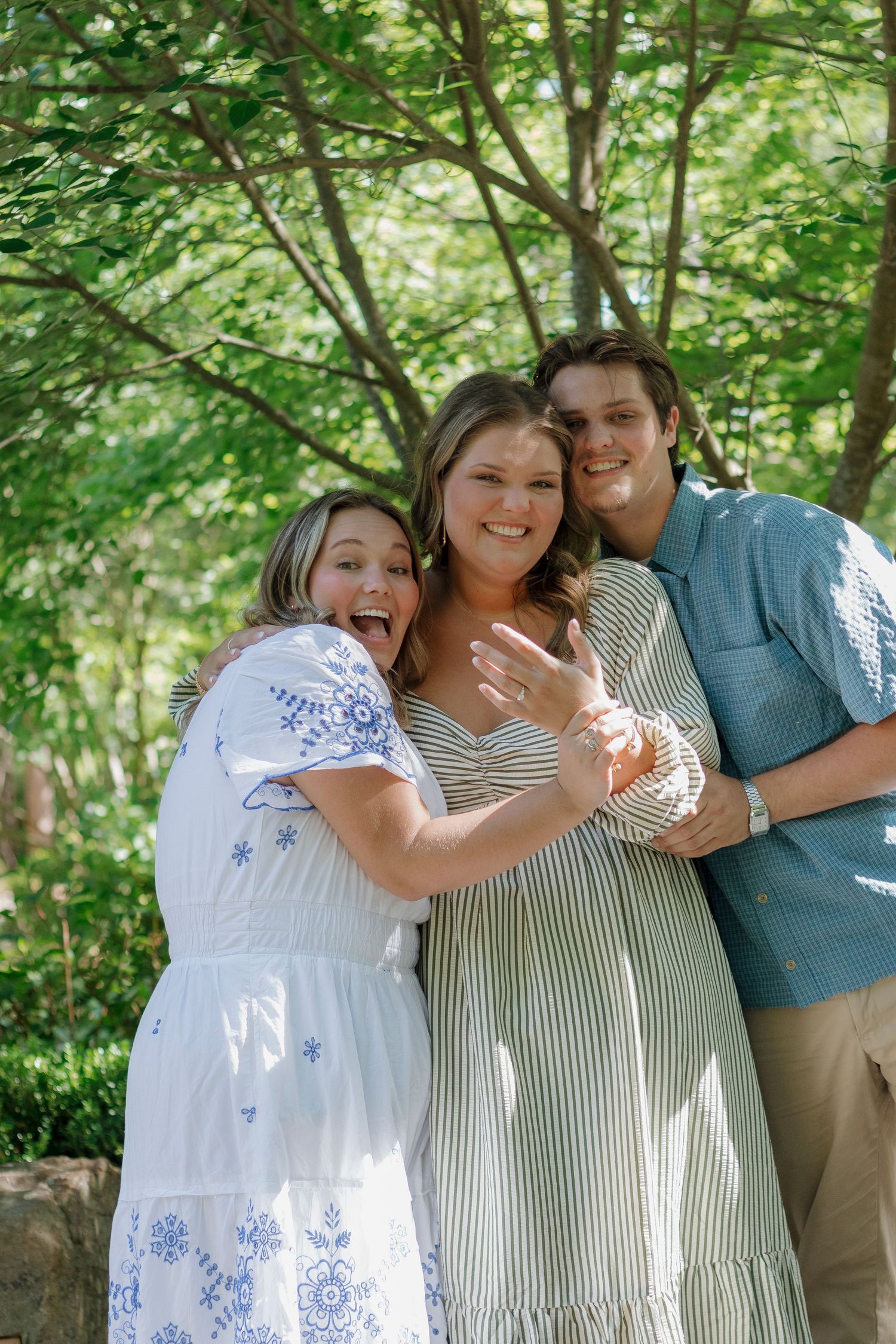 Three people smile outdoors, woman in center shows off engagement ring, surrounded by lush green trees.