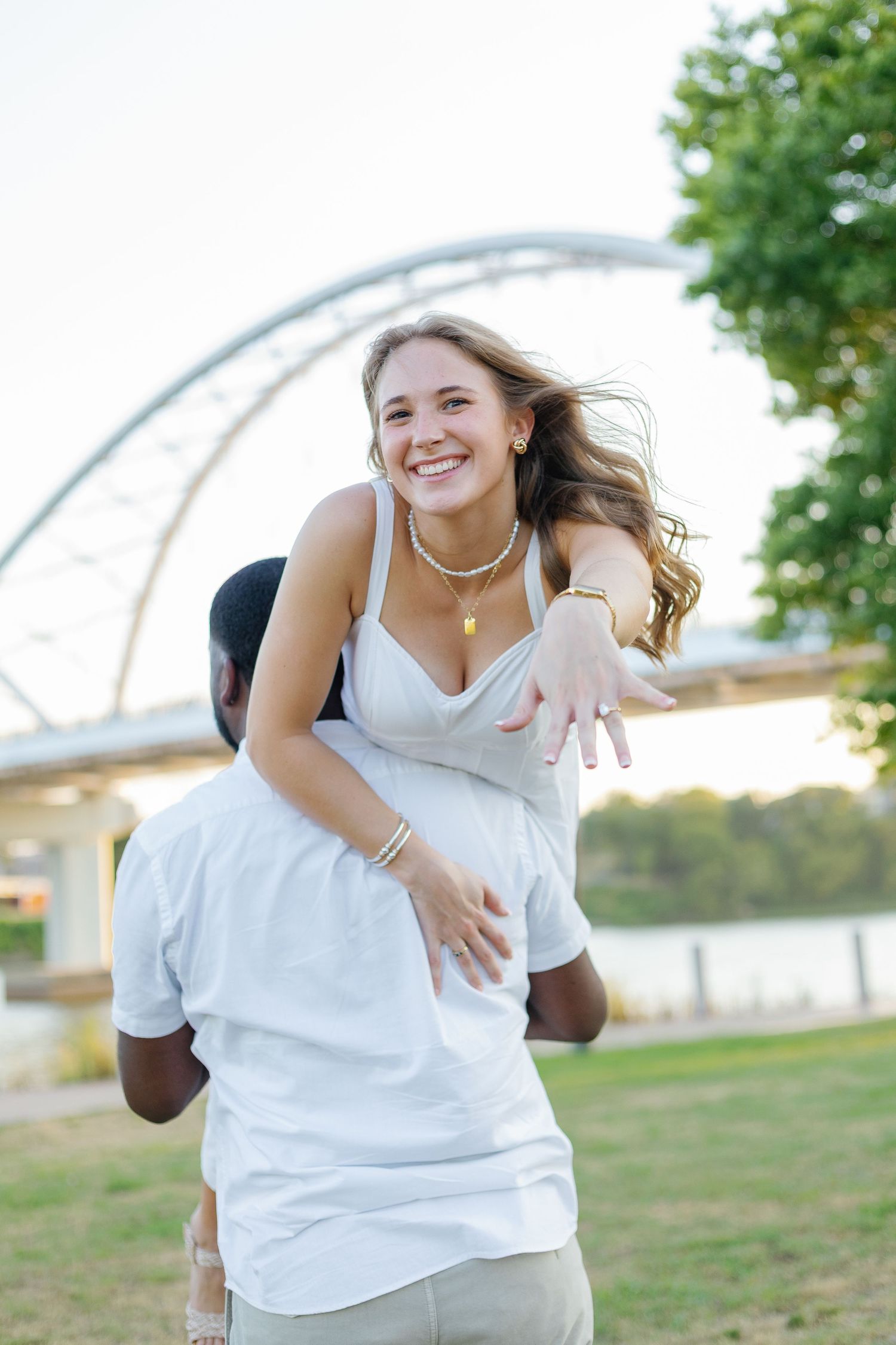 A smiling woman in a white dress gets a piggyback ride from a man outdoors near a bridge and river.