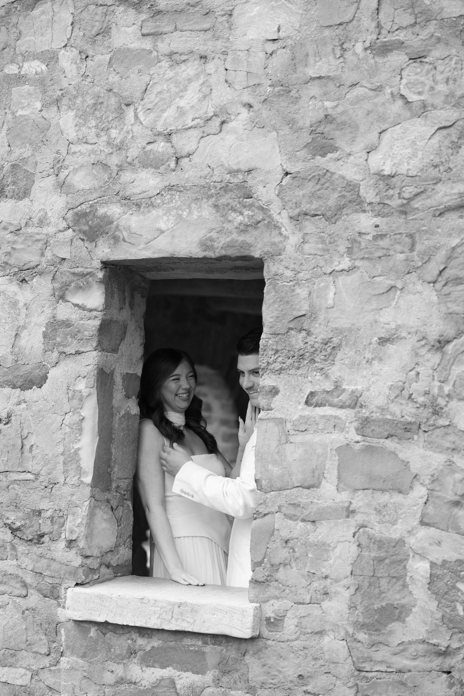 Black and white photo of couple smiling together through a stone window opening in a rustic wall.
