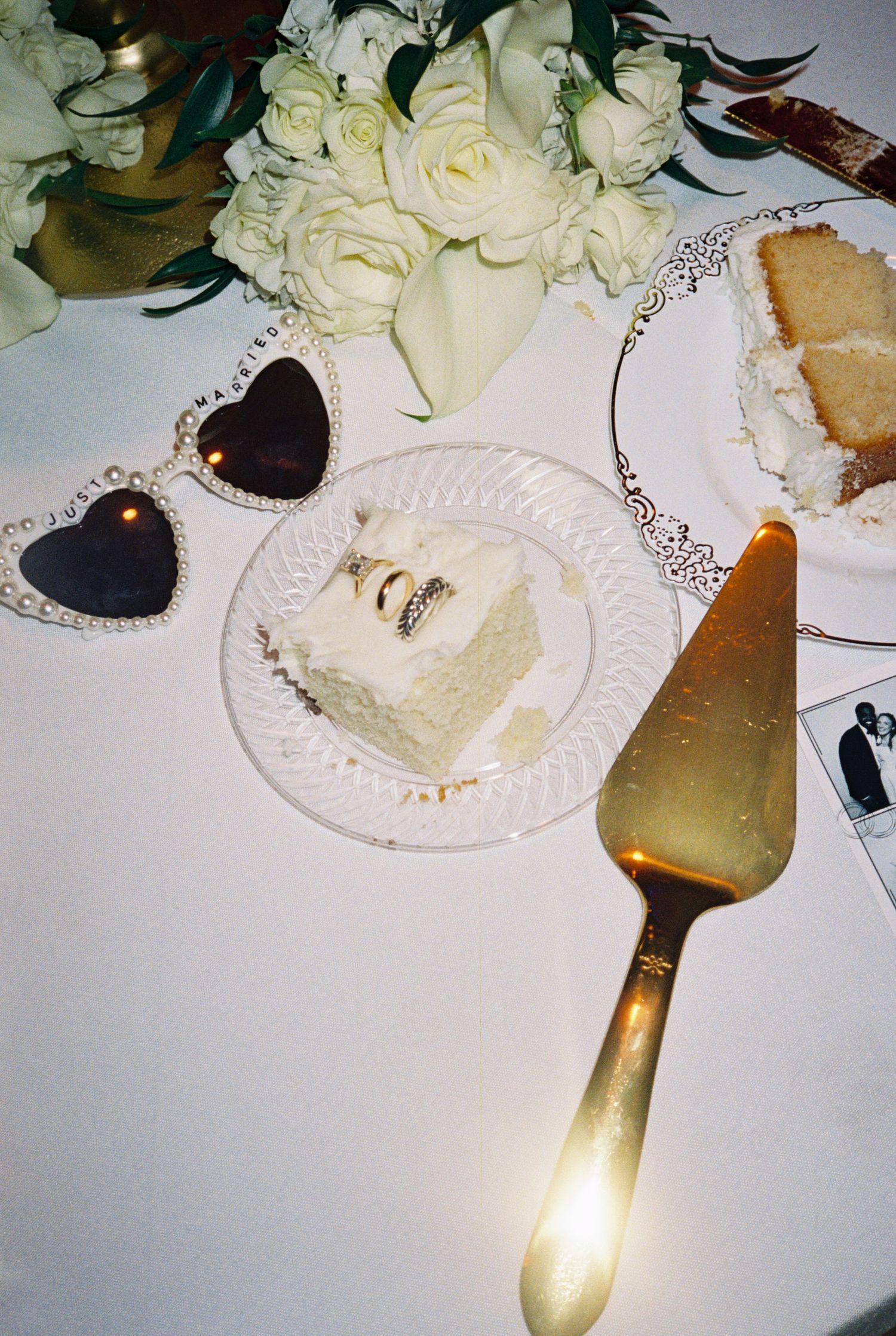 Slice of white wedding cake on crystal plate with gold server and heart sunglasses.