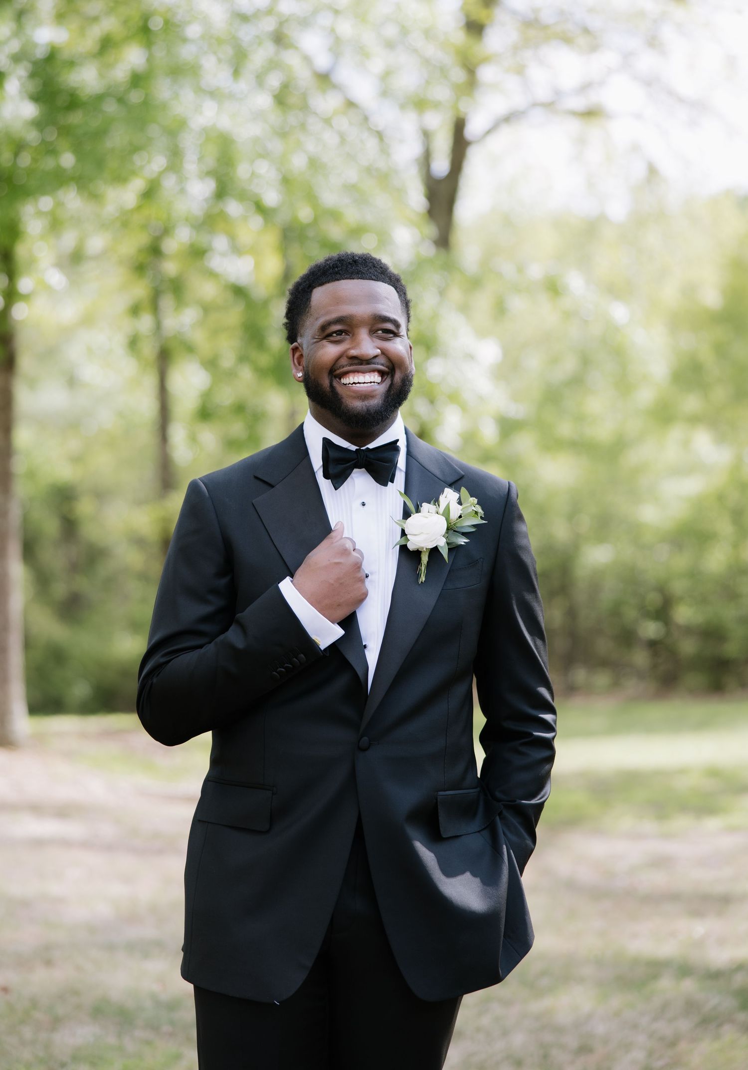 Dapper groom in black tuxedo adjusting bow tie with white floral boutonniere outdoors.