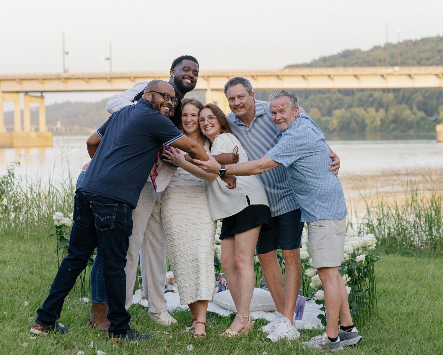 Group of six people hugging and smiling together outdoors near a river at sunset.