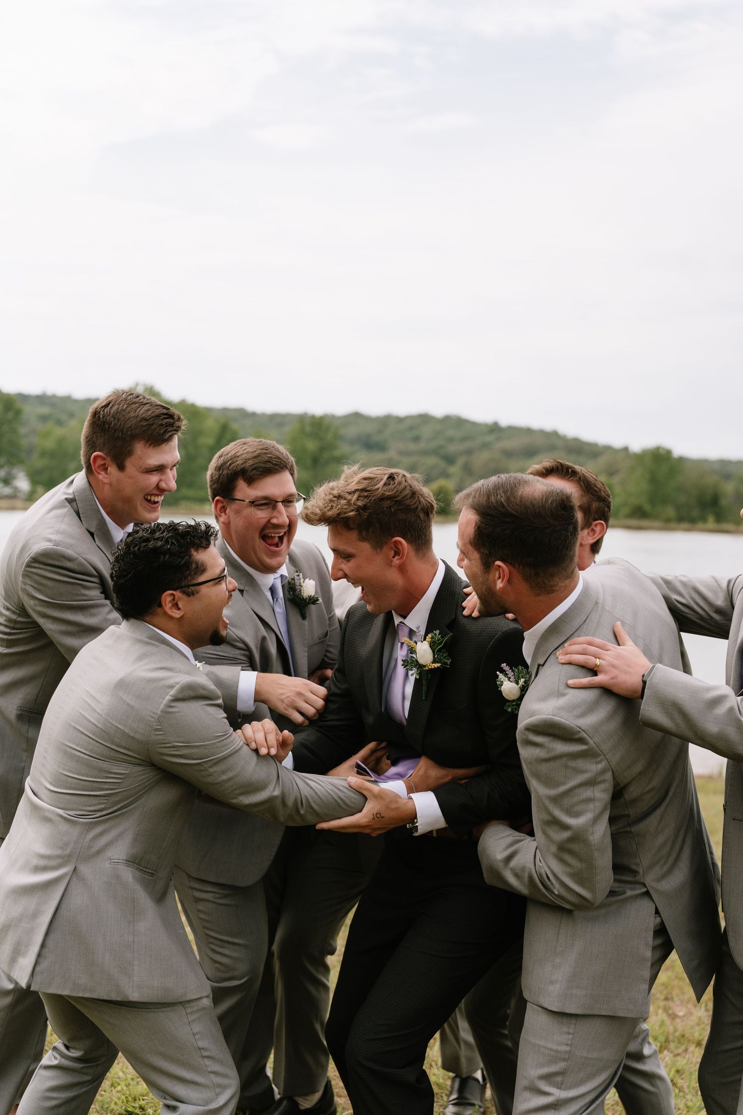 Groomsmen in gray suits laughing and surrounding groom in black tuxedo outdoors.