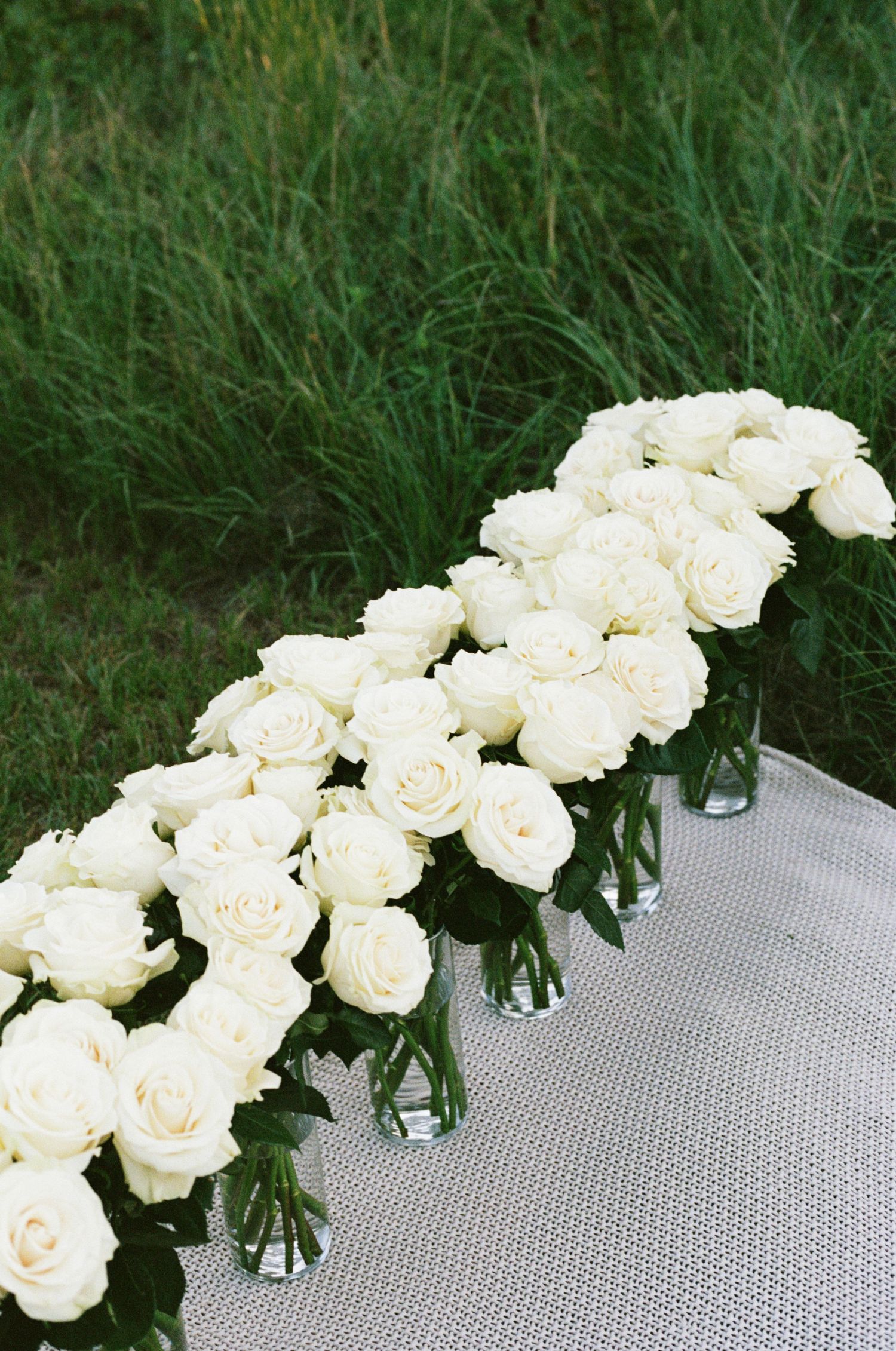 White roses arranged in glass vases on a silver table with green grass background, perfect for weddings.