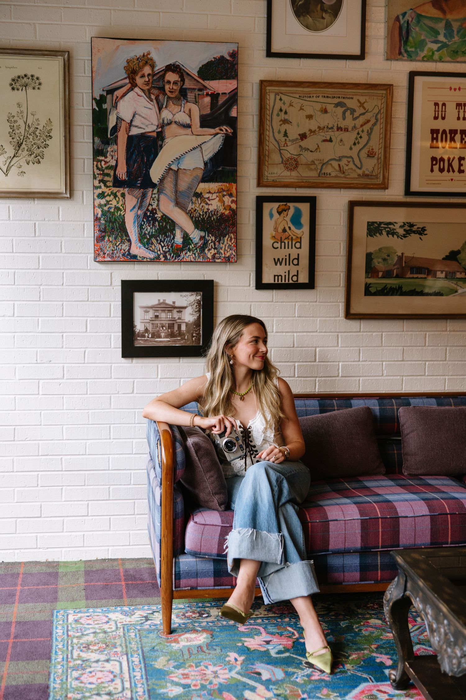 Woman sitting on plaid sofa in eclectic room with vintage gallery wall art and colorful floral rug.