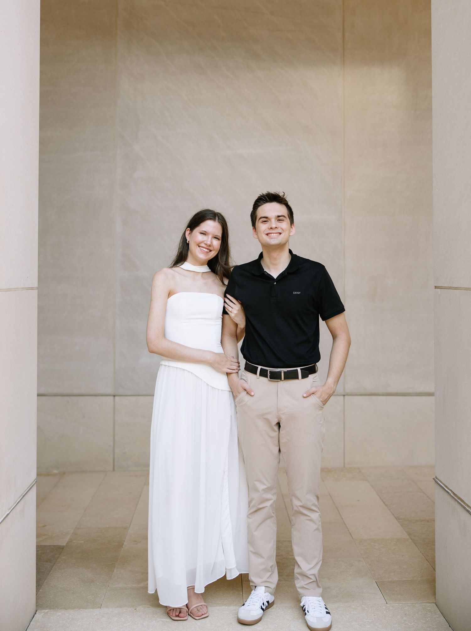 A smiling couple poses together, woman in white dress and man in black polo with khaki pants, against a beige stone wall.