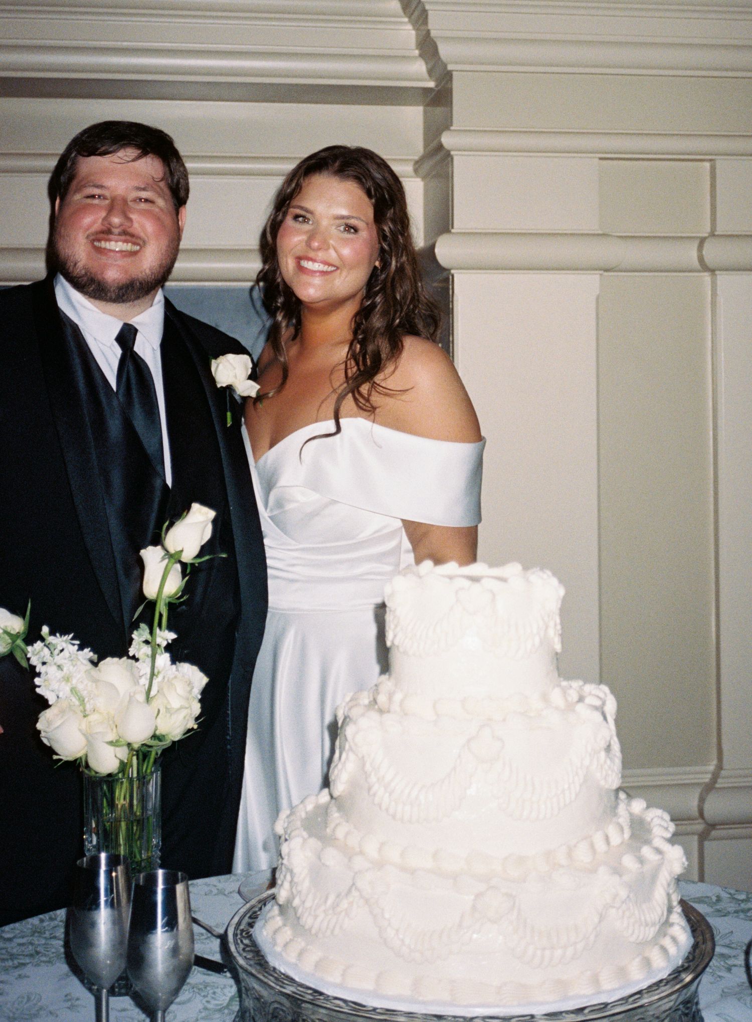 Bride and groom smiling beside a three-tier white wedding cake with floral decorations and roses in a vase nearby.