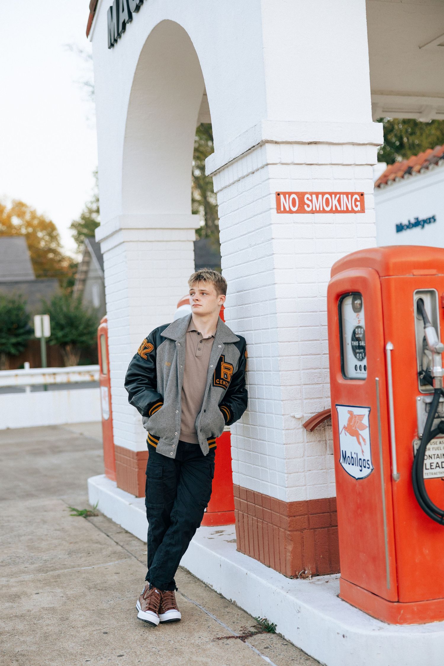 Young man in varsity jacket leaning against vintage Mobil gas station with No Smoking sign.