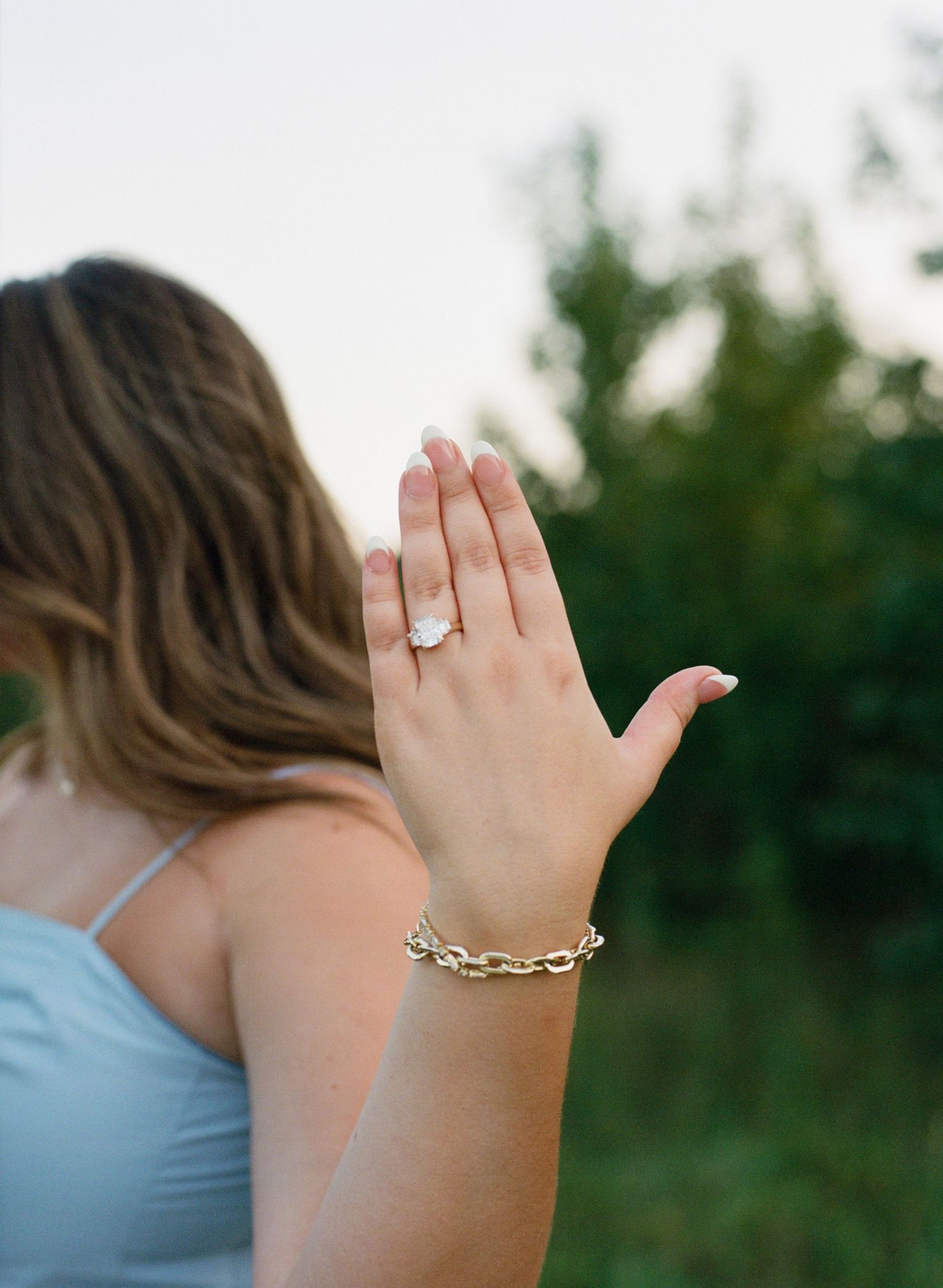 Woman showing off a large diamond engagement ring and gold chain bracelet with green trees in the background.