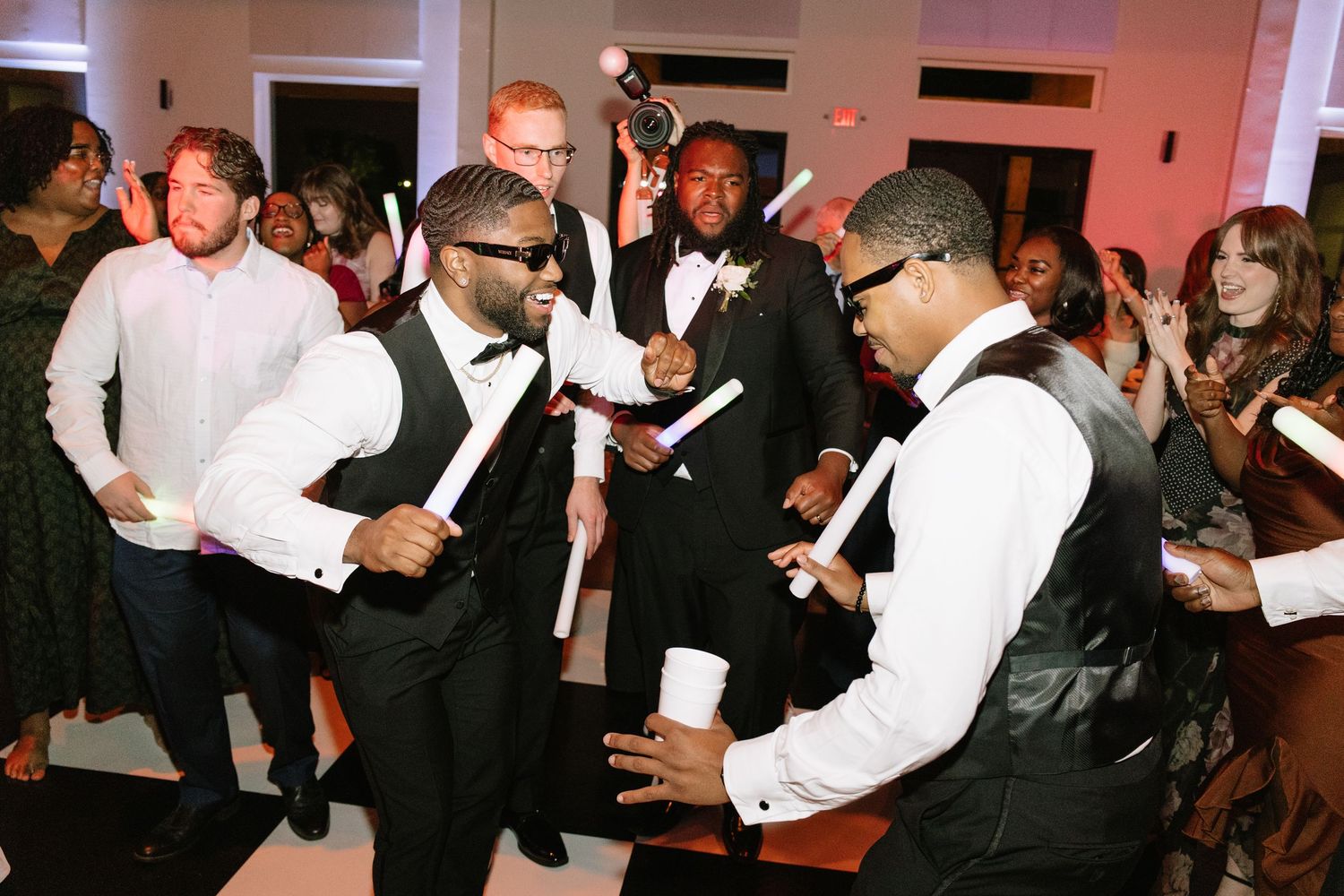 Two groomsmen in vests and sunglasses dance energetically at a wedding reception with guests in background.
