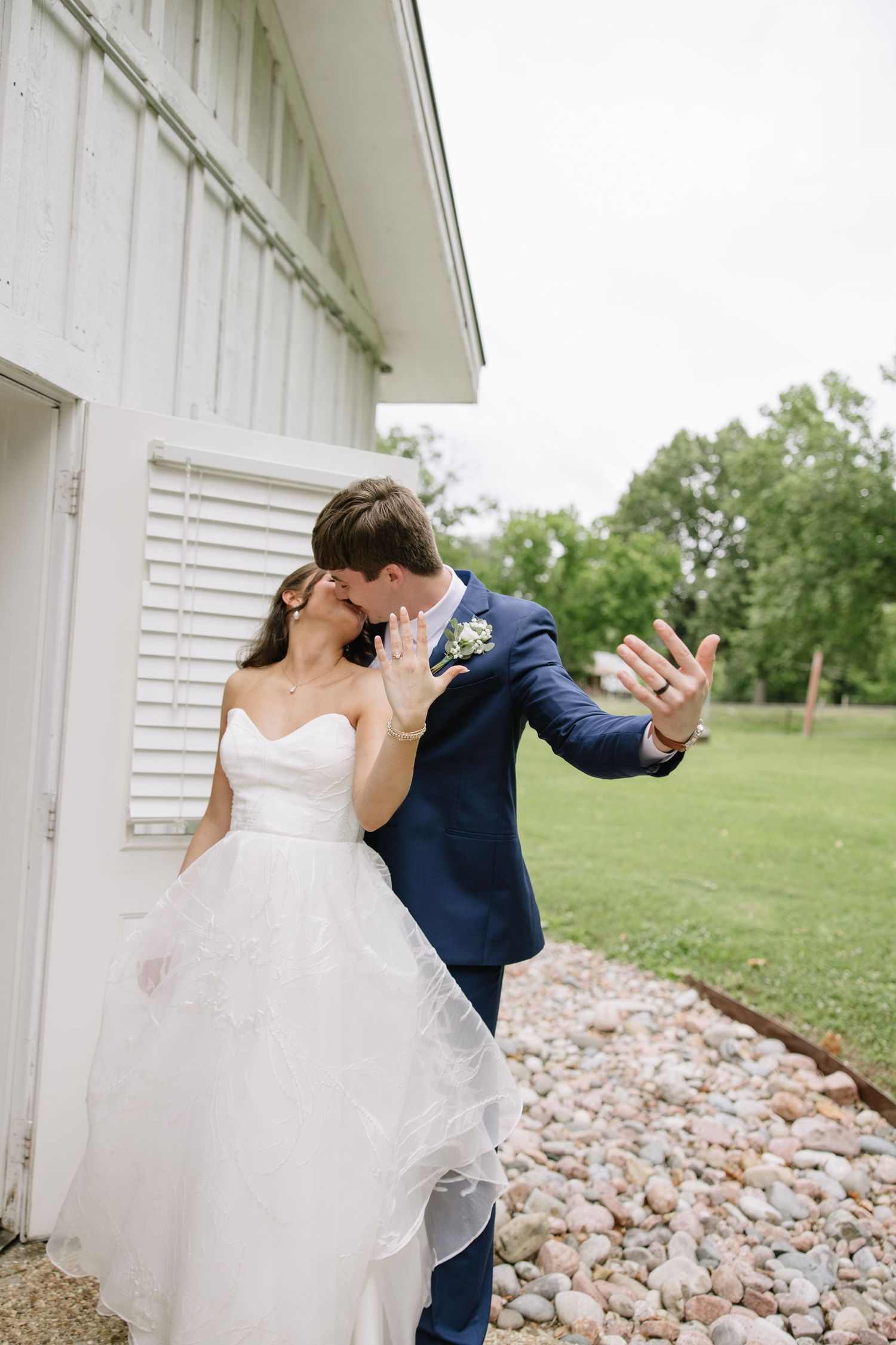 Bride and groom kissing beside white barn, showing off wedding rings, lush green trees in background.