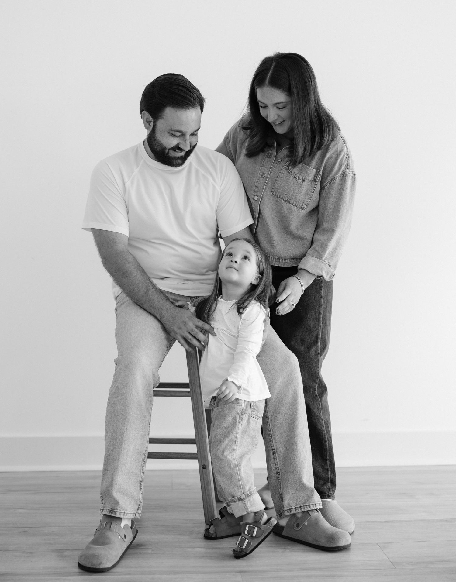 Black and white family portrait of parents and young daughter posing together on a stool in a studio.