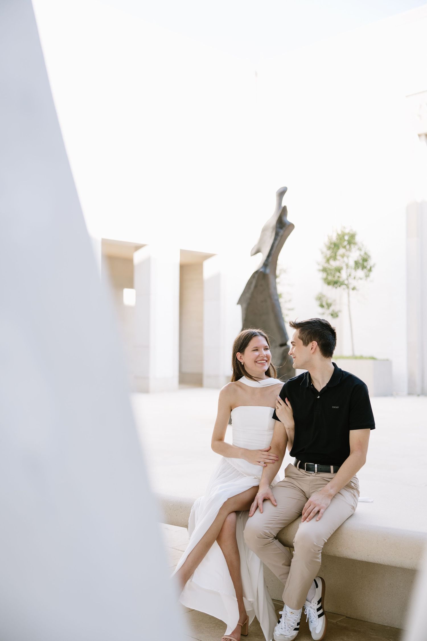 A couple sits closely on a white ledge, smiling at each other, with a bronze sculpture and modern architecture in the background.