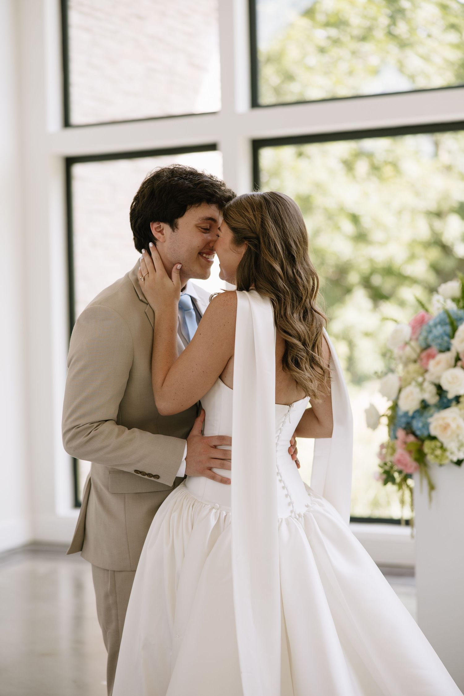 A bride and groom share a romantic almost-kiss moment, dressed in a white gown and beige suit near large windows.