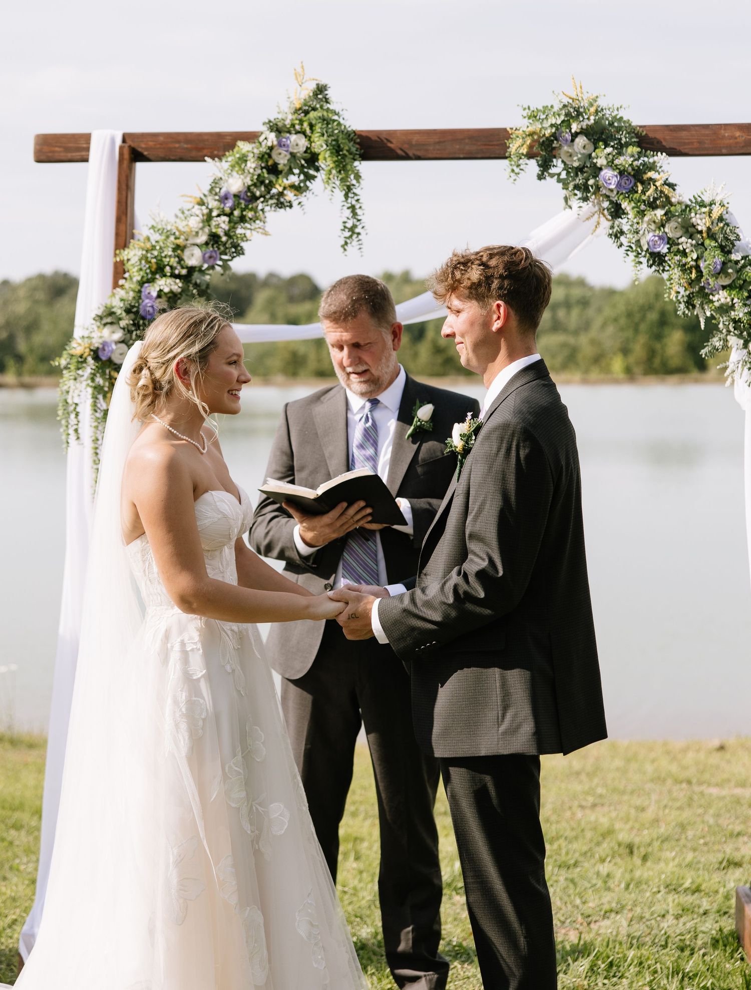 Bride and groom exchange vows outdoors under a floral arch by a lake, officiant holding a book.