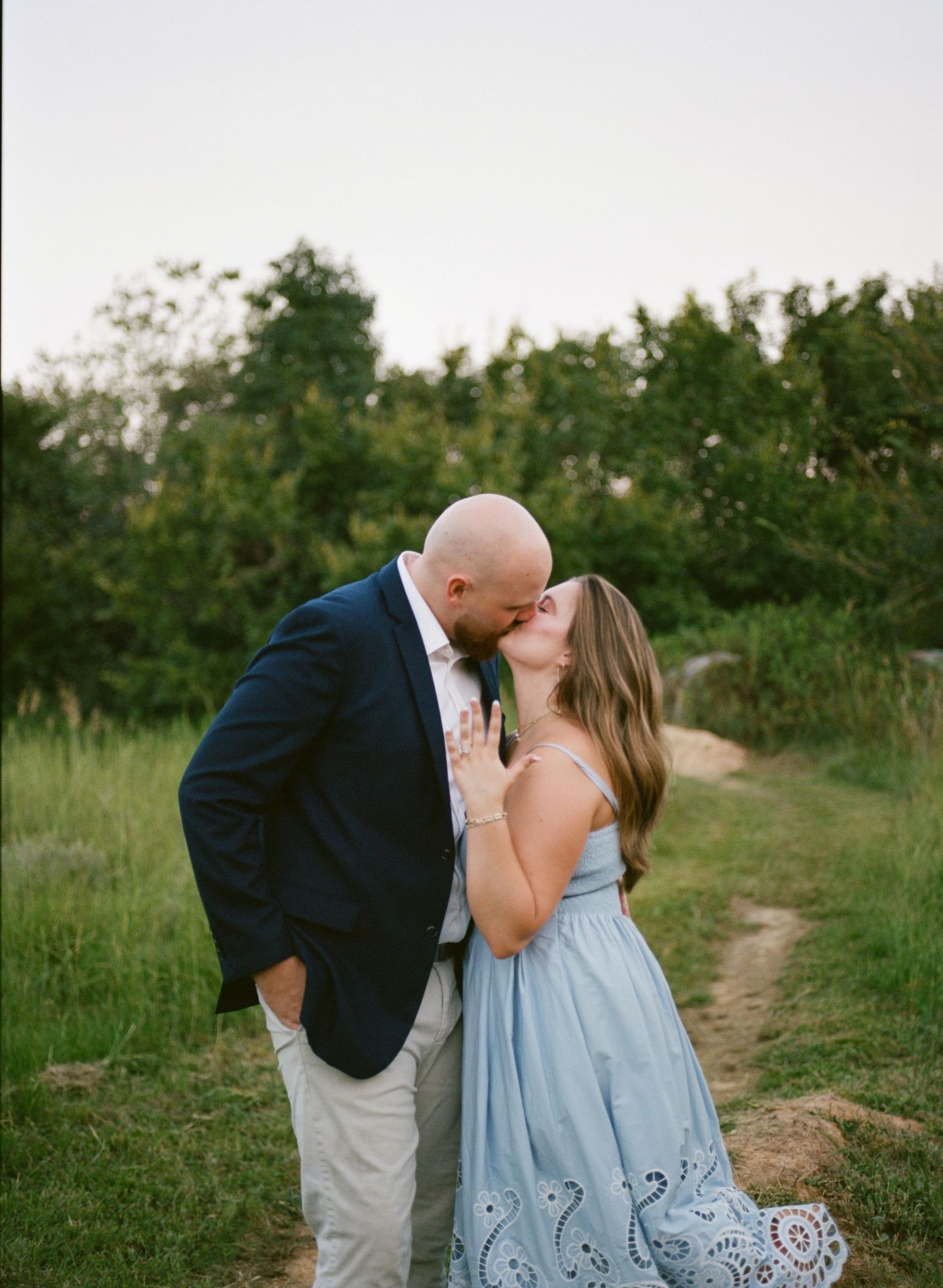 Couple sharing a romantic kiss outdoors on a dirt path surrounded by lush green trees and grass at dusk.