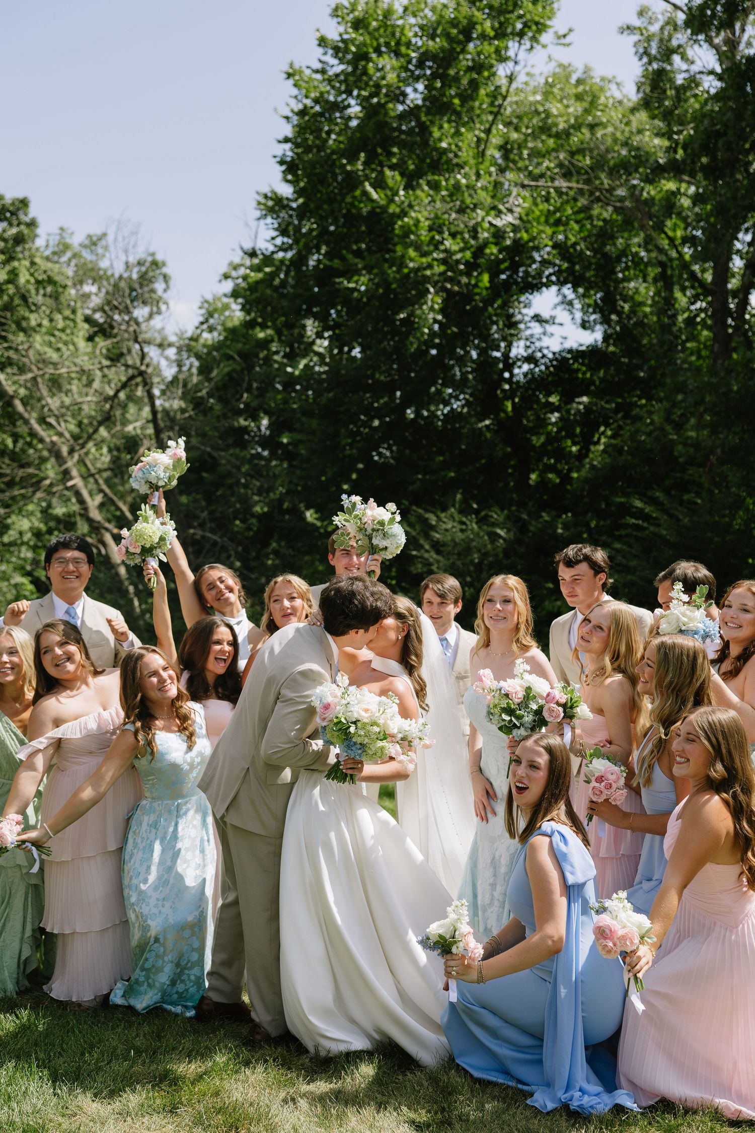 Bride and groom share a kiss surrounded by cheerful wedding party in pastel dresses outdoors.