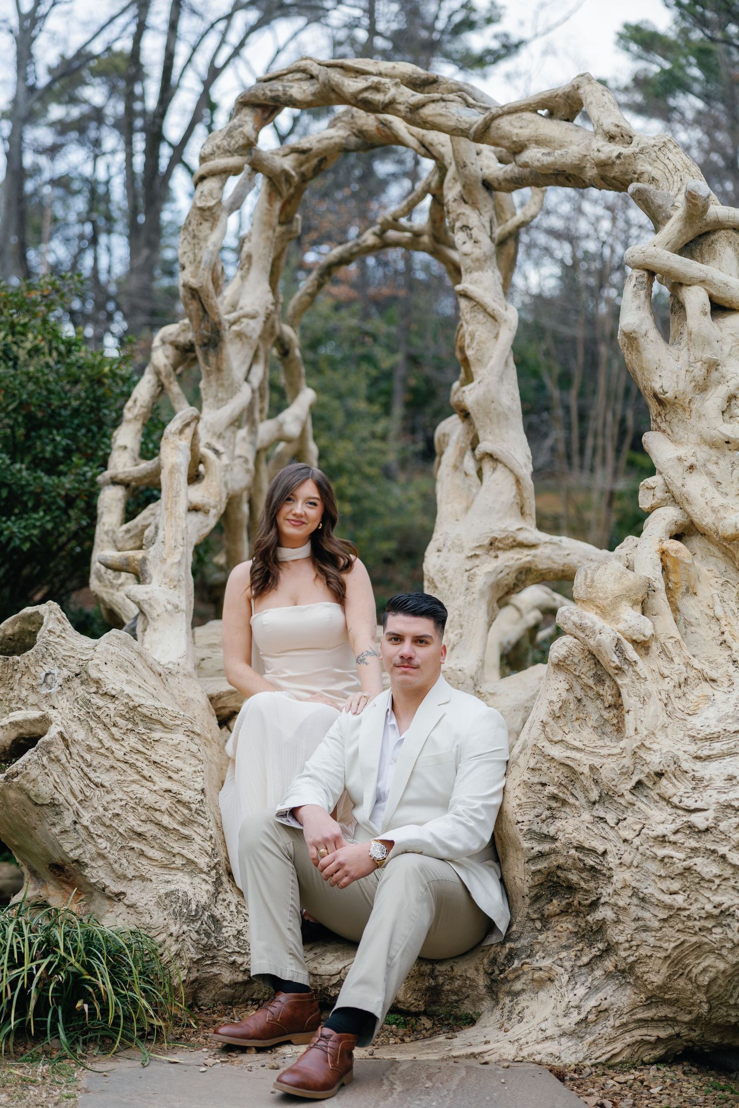 Couple posing on a rustic stone arch sculpture in a wooded garden, woman seated above man in elegant attire.