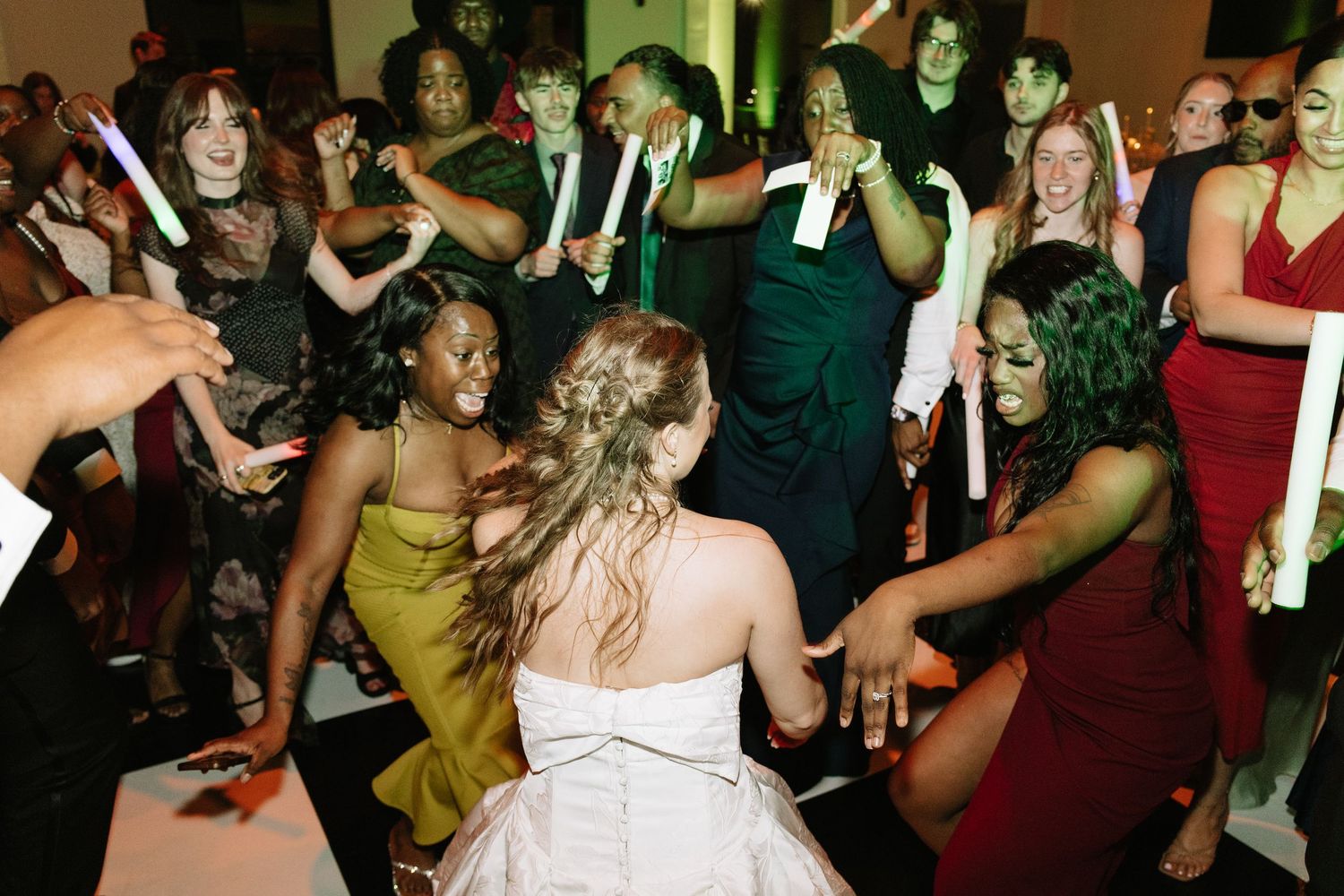 Bride in white gown dancing with guests holding glow sticks at a vibrant wedding reception celebration.