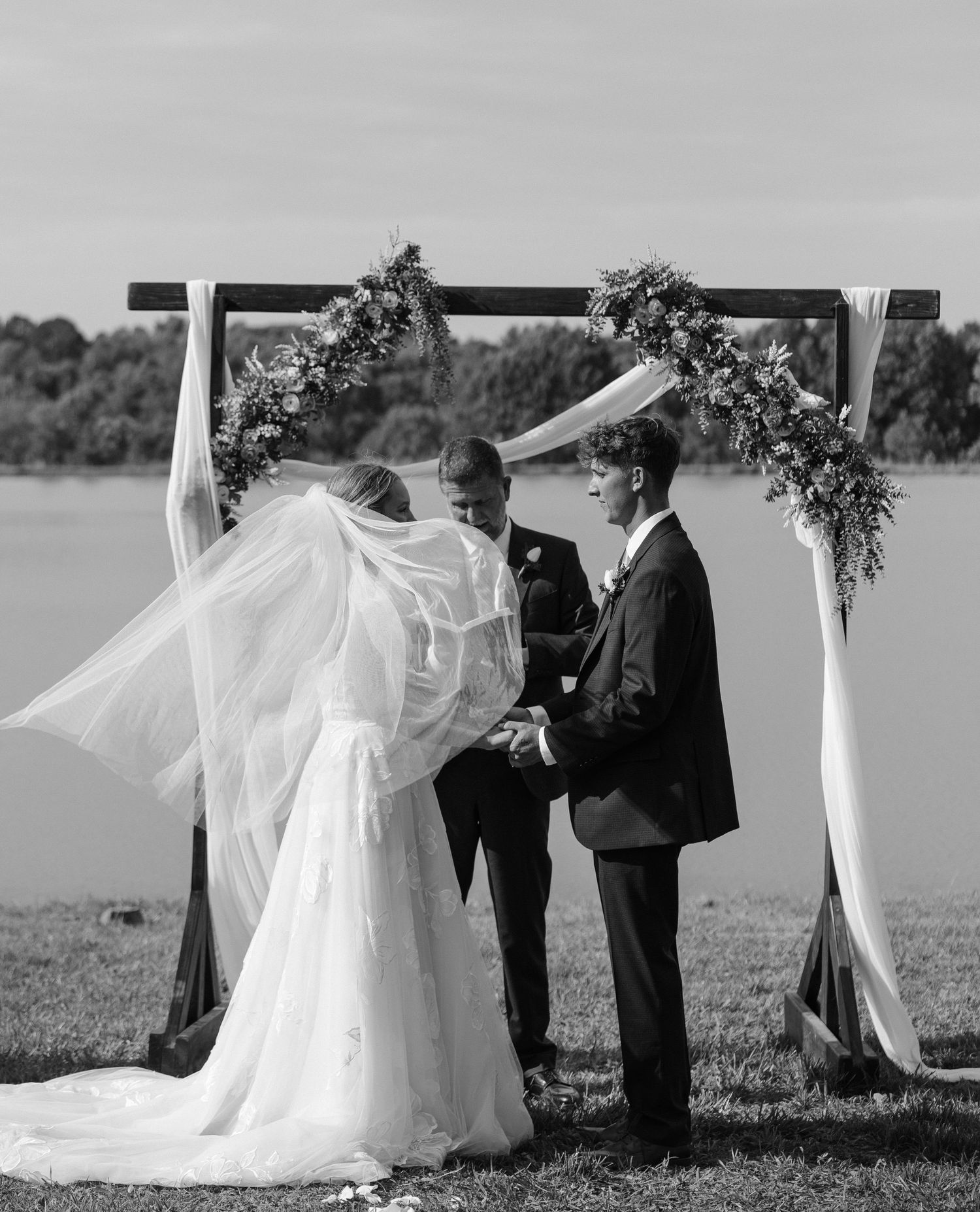 Black and white photo of a bride and groom exchanging vows under a floral arch by a lake.