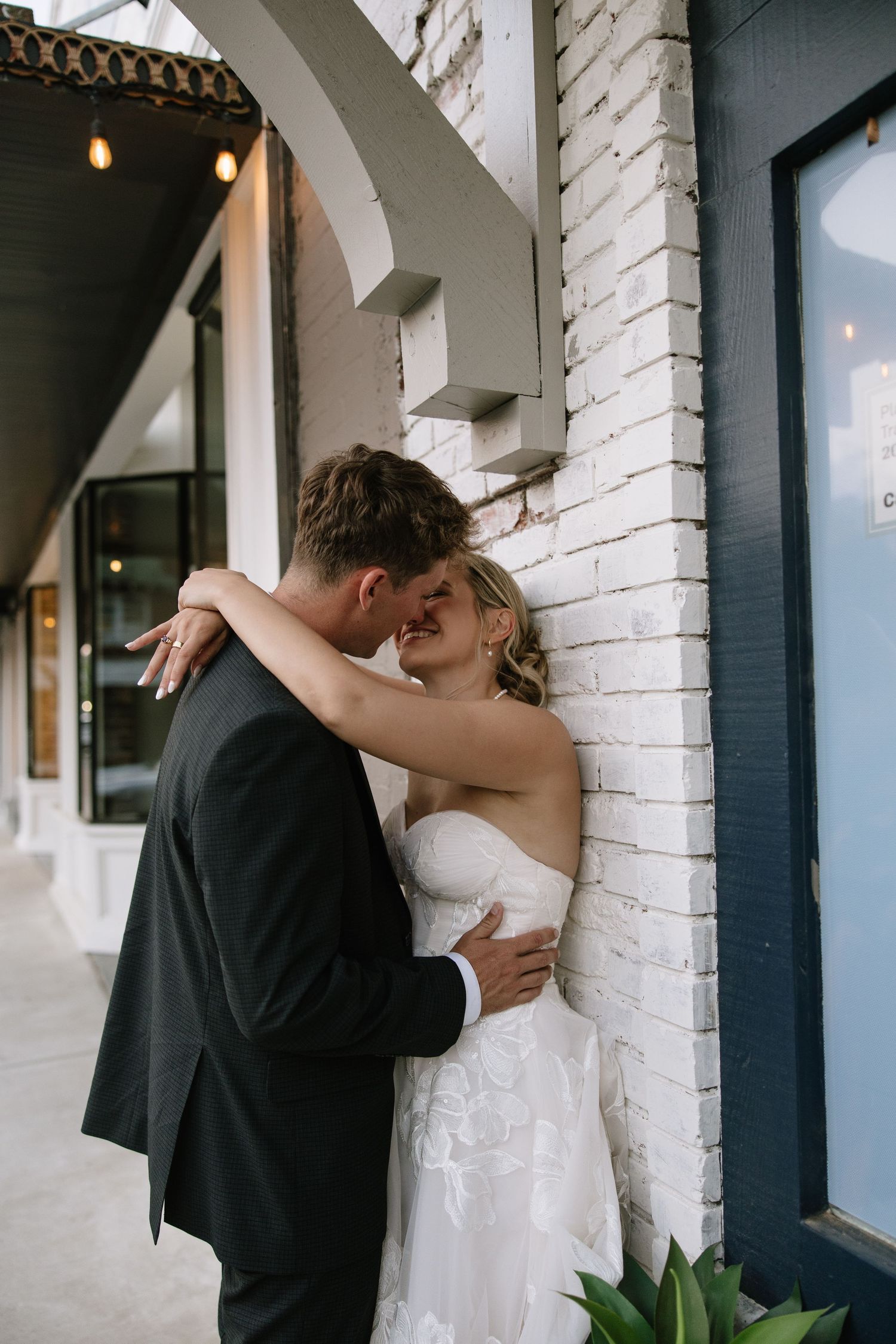 Bride and groom share a romantic moment leaning against a white brick wall outdoors in wedding attire.