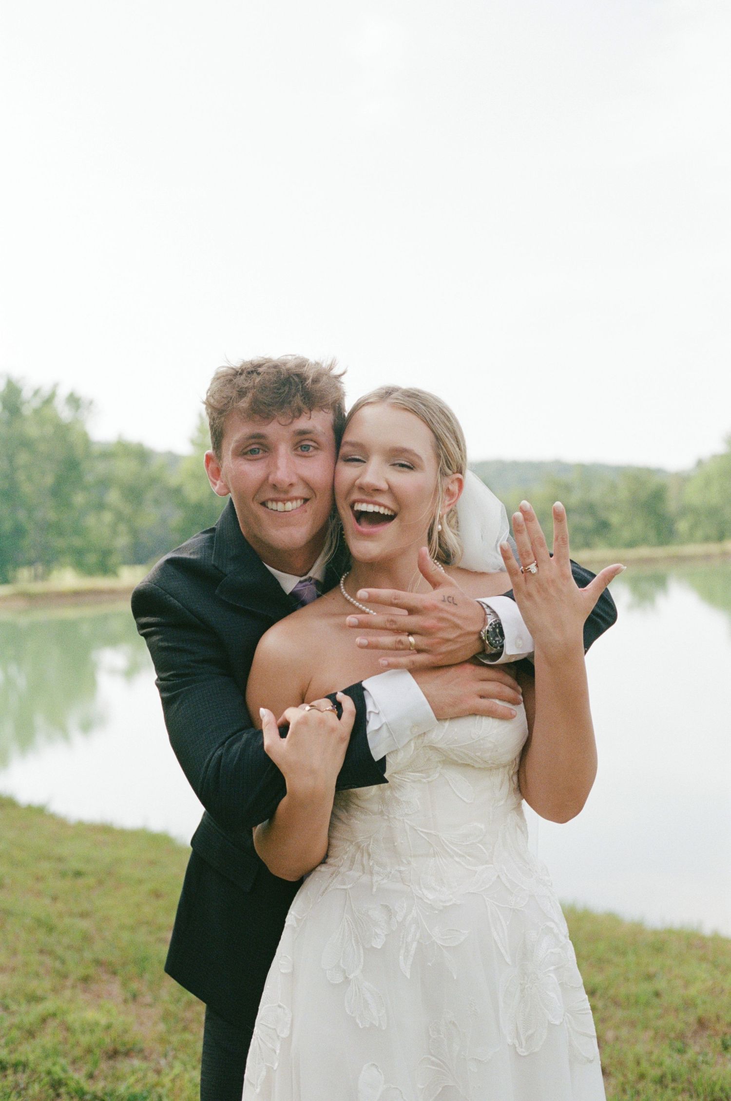 Happy newlyweds embrace outdoors by a lake, bride in white gown showing wedding ring, groom in black suit smiling.