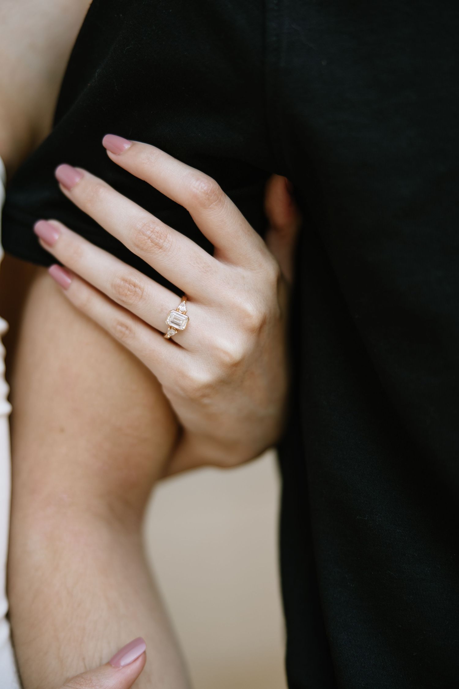 Close-up of a woman's hand wearing a diamond engagement ring on gold band, resting on a man's arm.