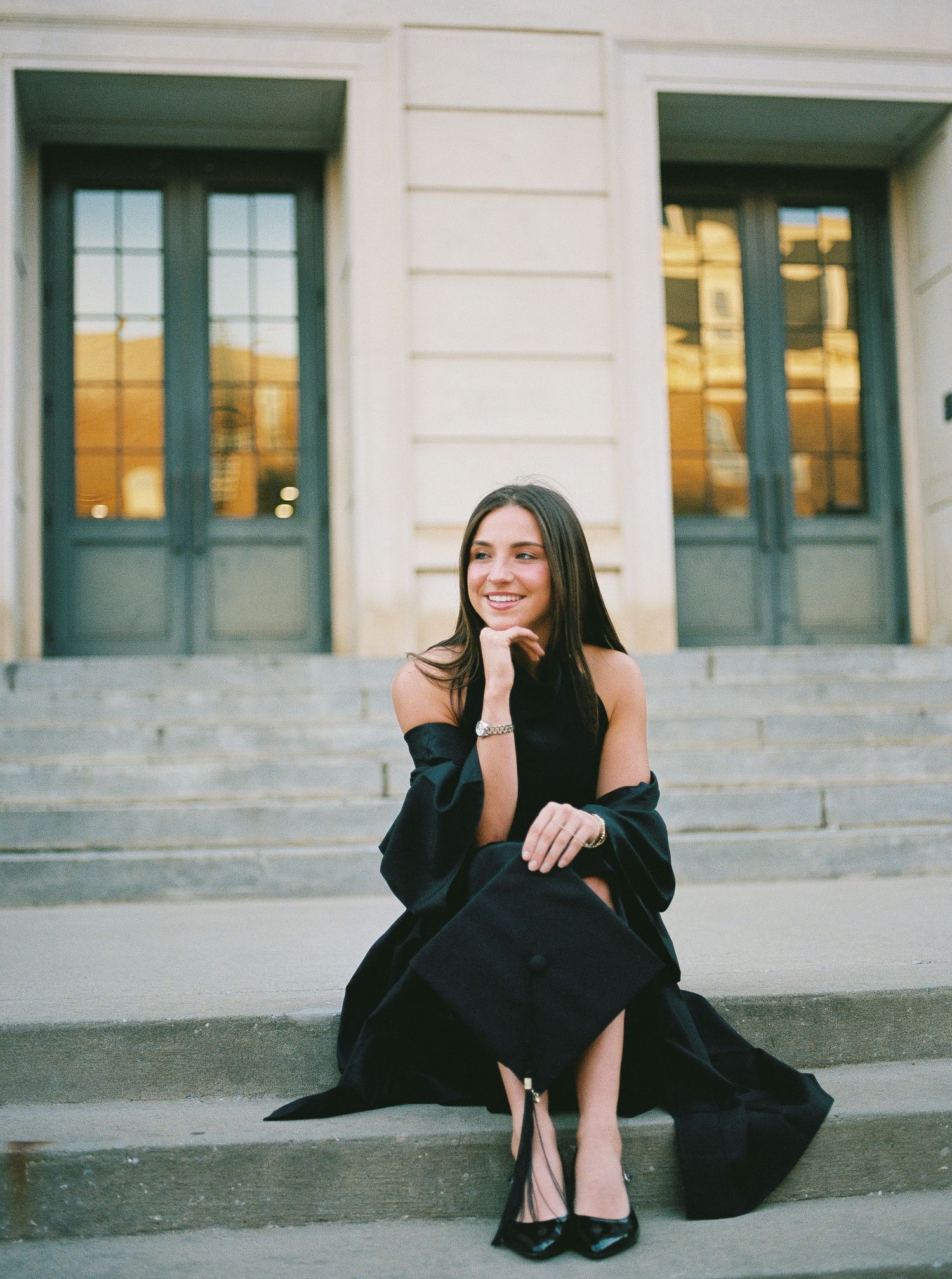 Elegant graduate in black dress and gown sitting on steps of a classic building with tall doors.