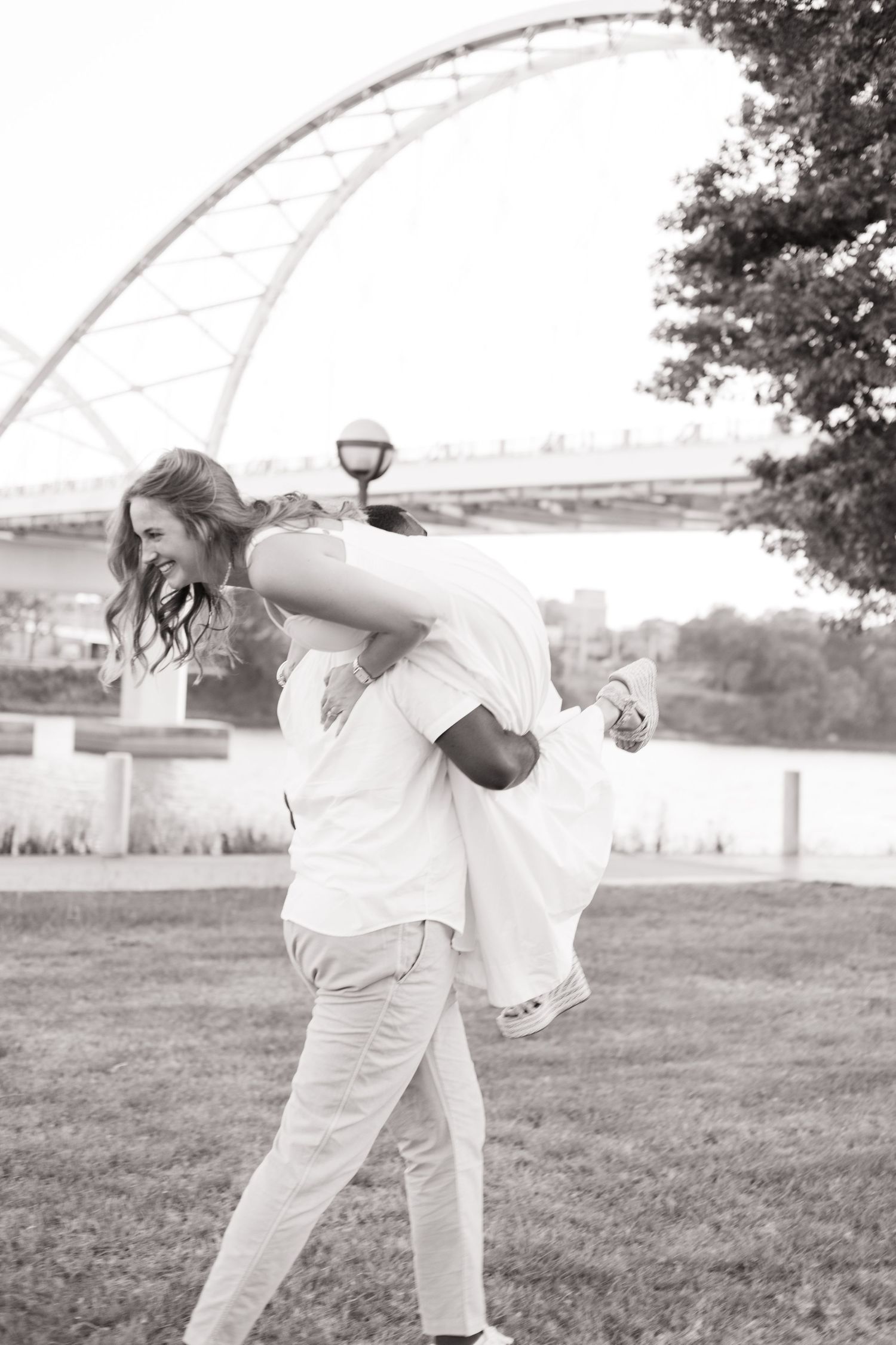 Black and white photo of a man carrying a laughing woman on his back in a park near a bridge.