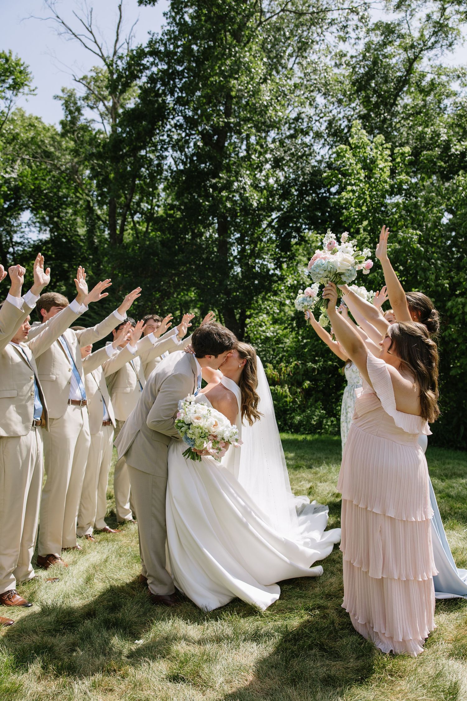 Bride and groom share a kiss while wedding party cheers with raised arms outdoors on a sunny day.