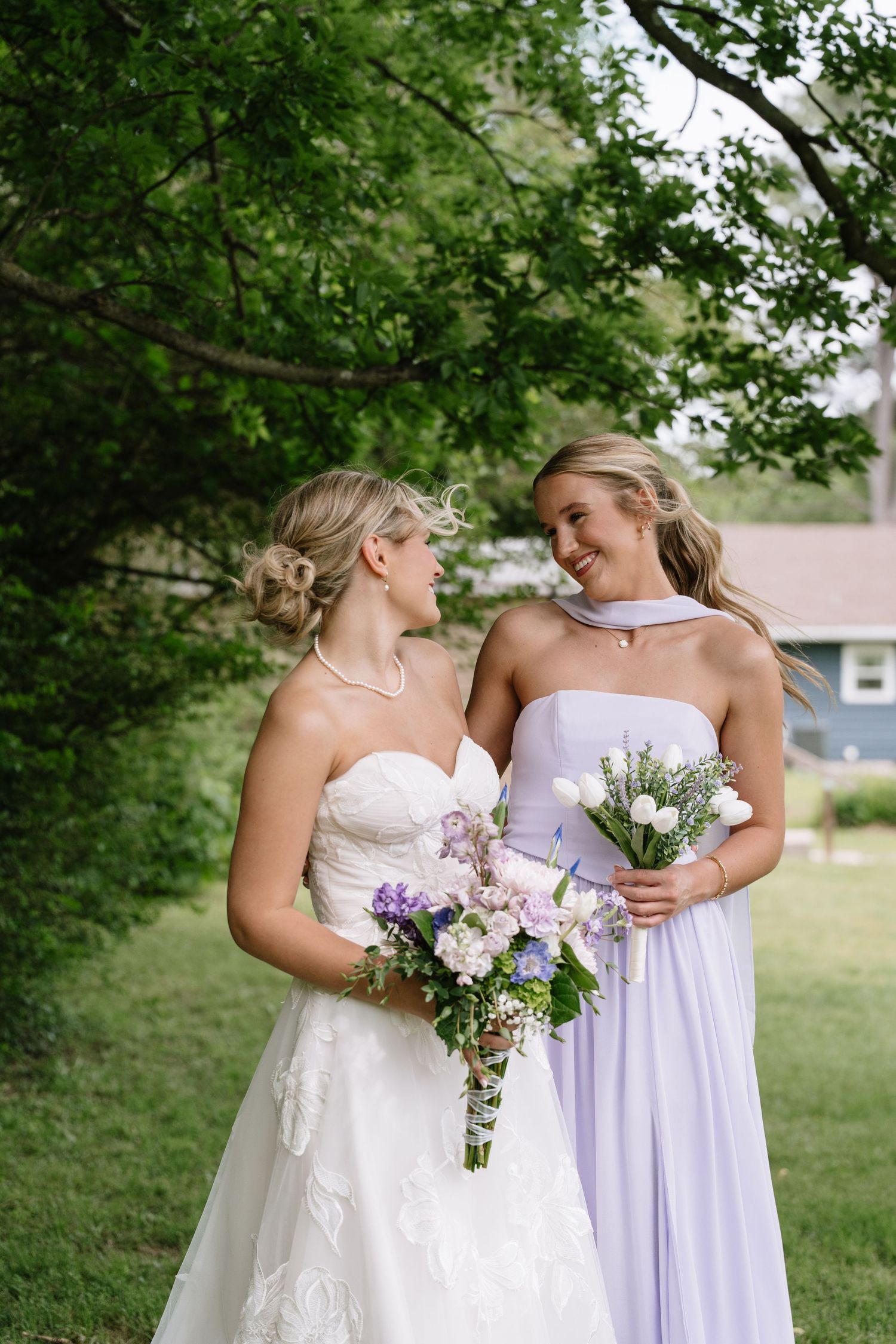 Bride in white lace gown and bridesmaid in lavender dress smiling while holding bouquets outdoors.