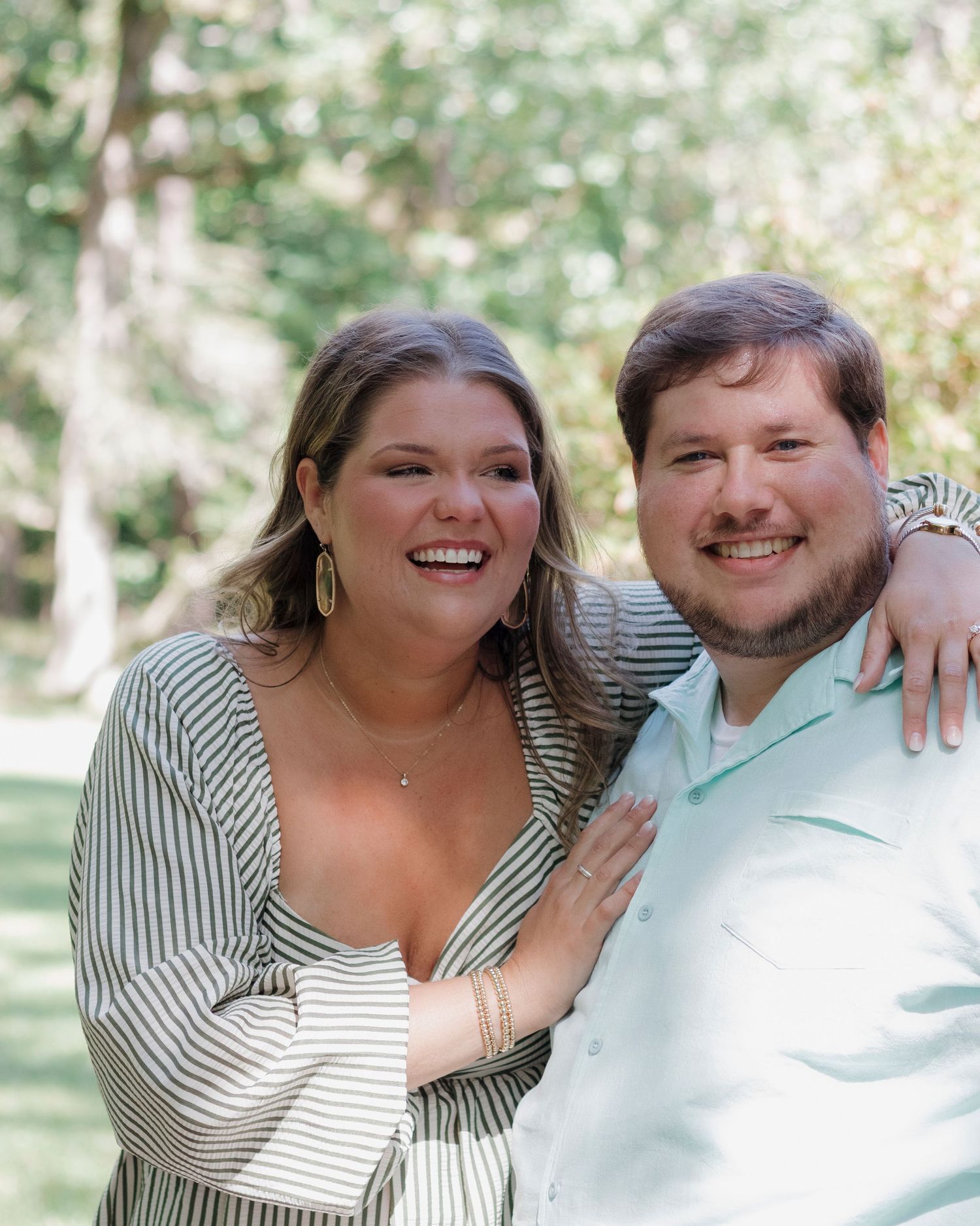 Smiling couple posing outdoors in a lush green park, woman in striped dress with man in mint shirt.