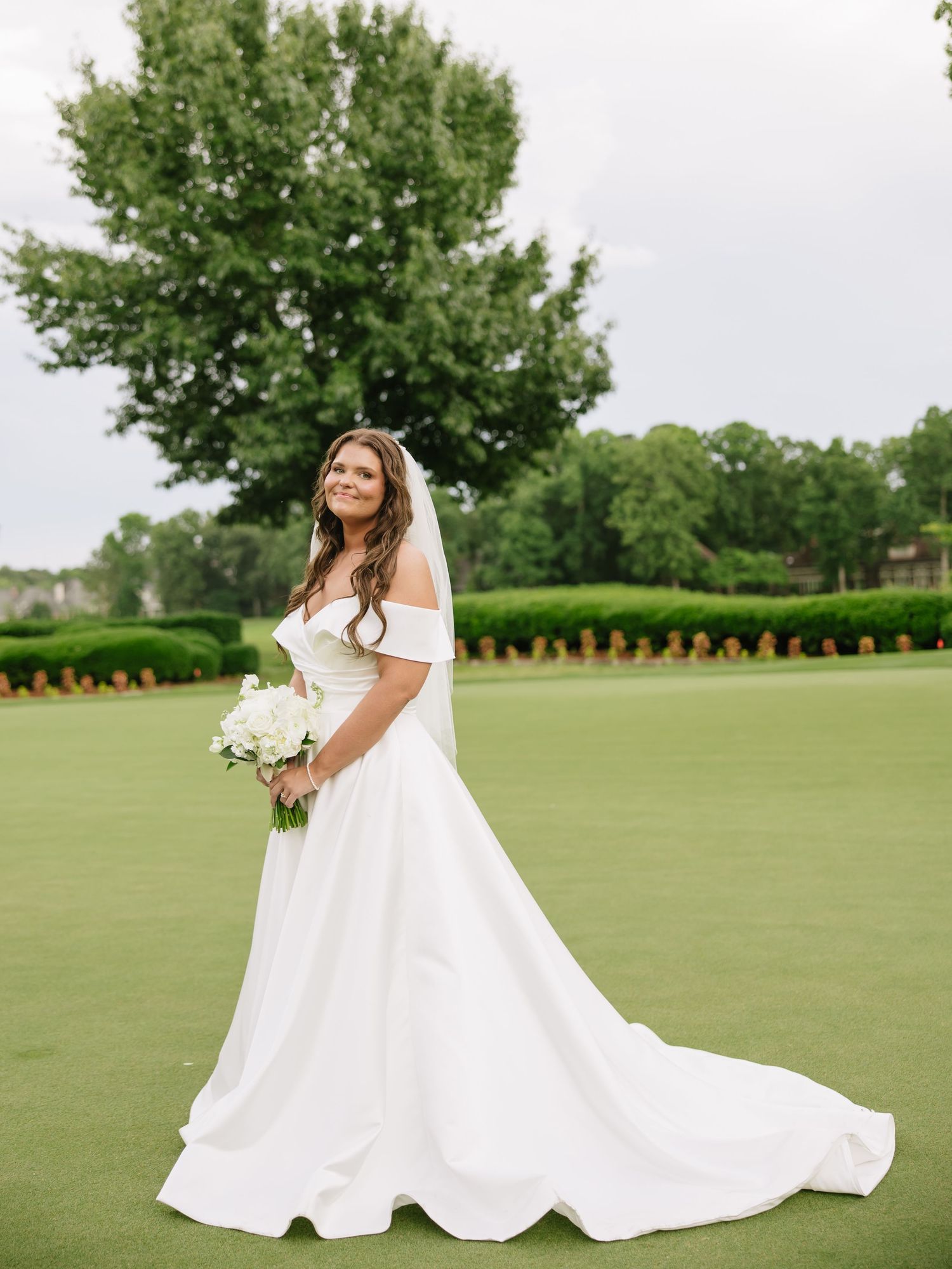 Bride in off-shoulder white gown with train holds bouquet on green golf course lawn.