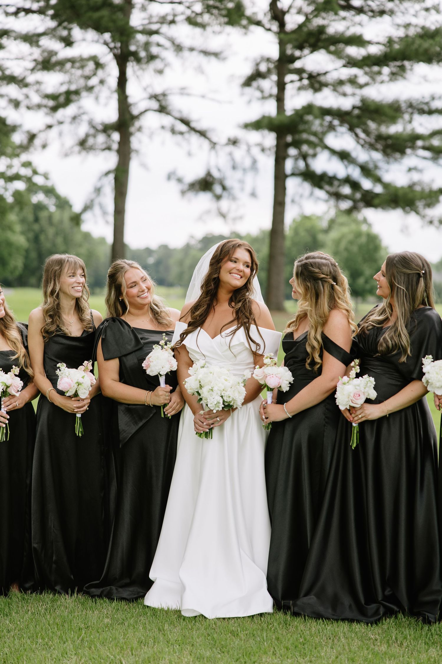 Bride in white off-shoulder gown with bridesmaids in black dresses holding white bouquets outdoors.