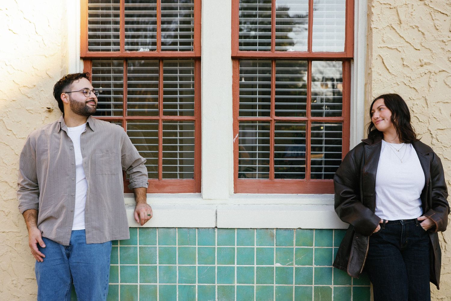 Two people standing apart against a teal tiled wall with windows, smiling outdoors.
