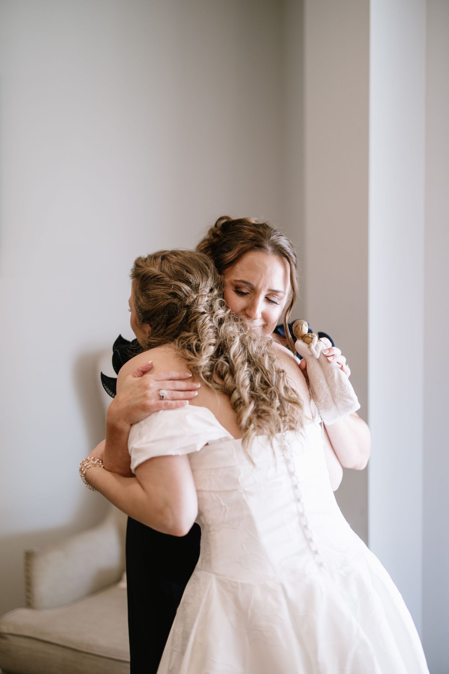Emotional bride and mother sharing a tender hug during wedding day preparations in a softly lit white room.
