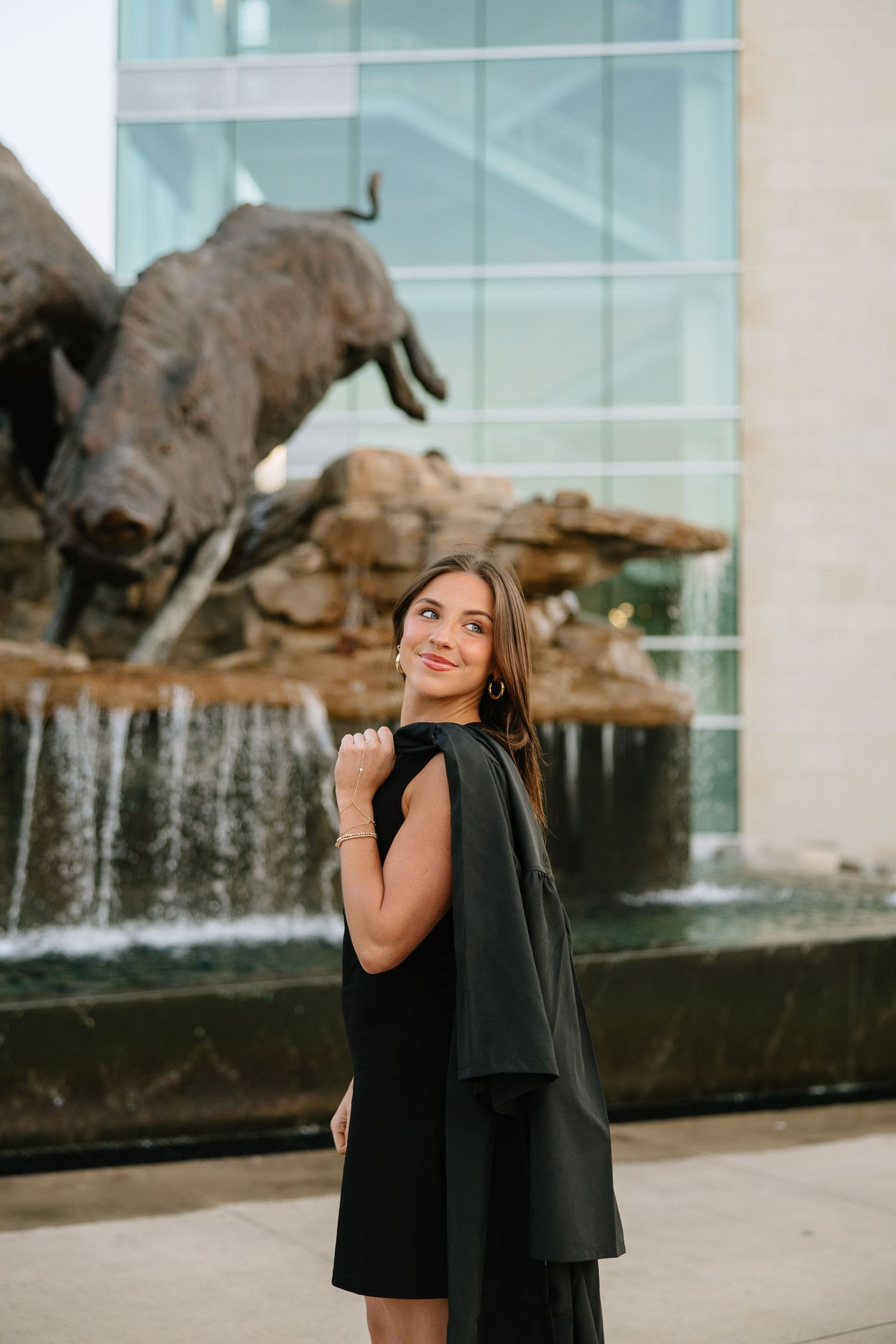 Elegant woman in black cape dress poses before bronze bull sculpture and glass building fountain.
