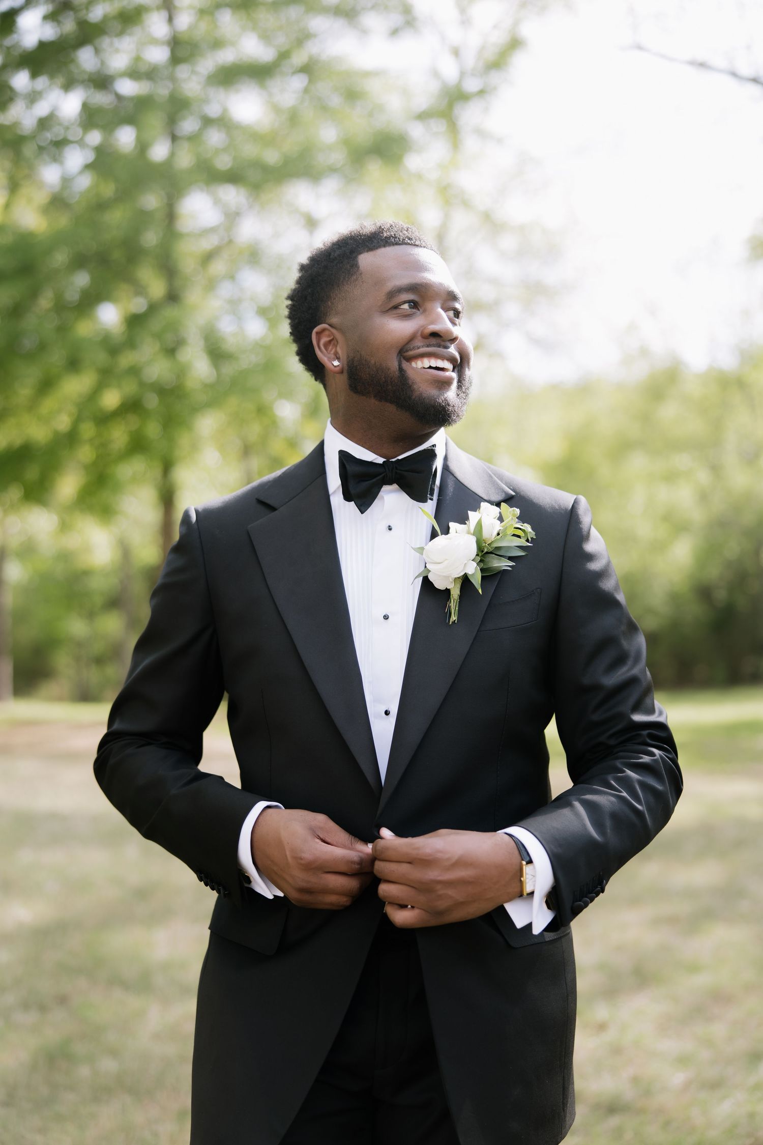 Smiling groom in black tuxedo with white boutonniere buttoning jacket outdoors.